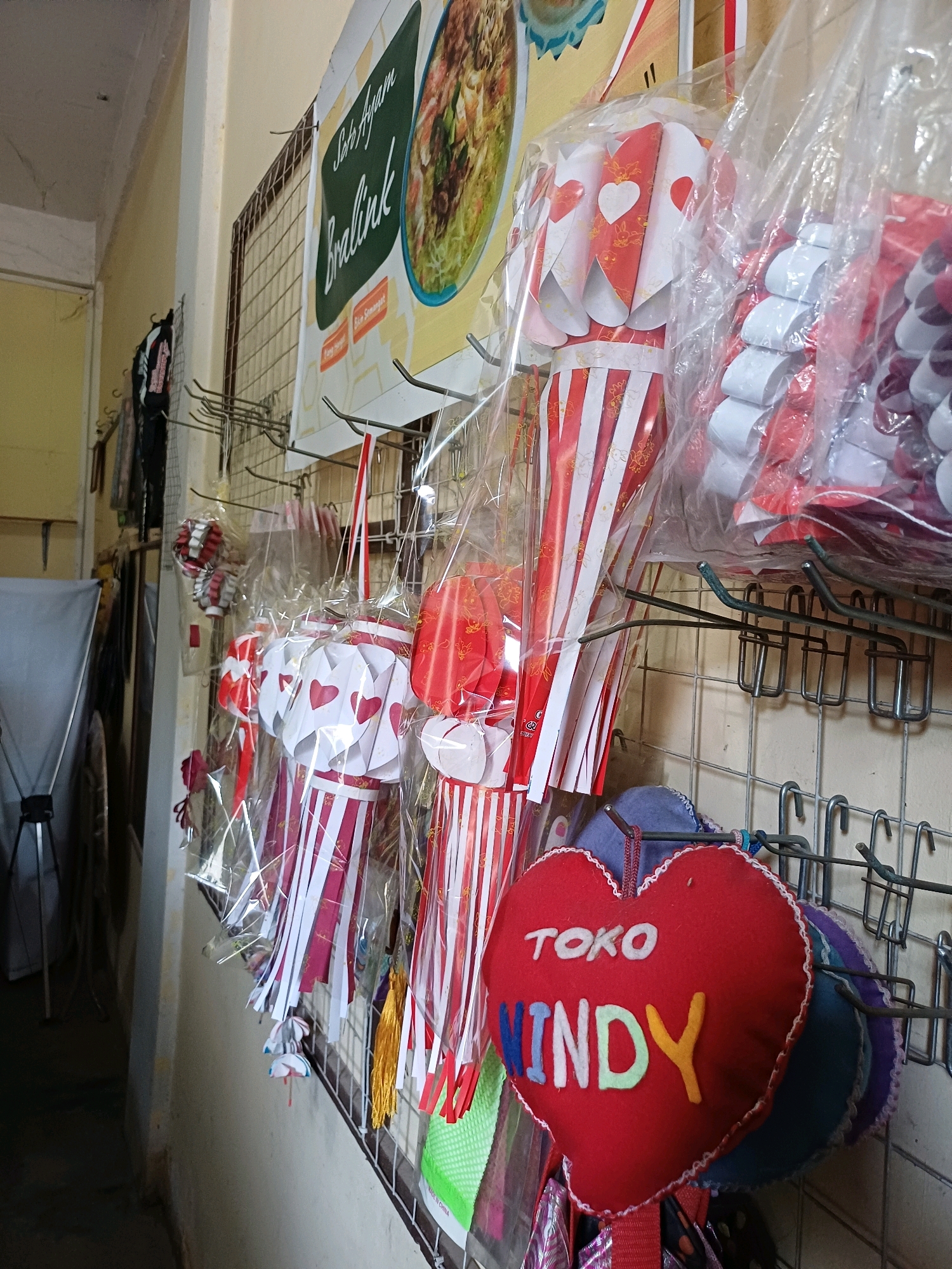 The image shows a wall display of various decorative items, likely for sale. The wall is a light beige color, and a wire grid hangs on it, holding the merchandise. In the foreground, a red heart-shaped pillow is prominently displayed, with the text "TOKO MINDY" written on it in colorful letters. Behind the pillow, several other heart-shaped pillows of different colors and layered fabric are visible.
Hanging from the wire grid are numerous colorful paper lanterns or decorations. Many of these are red and white, with striped patterns and heart cutouts. Some are enclosed in clear plastic packaging. To the left of the lanterns, a sign with text and an image of food is partially visible. The overall impression is that of a small shop or stall selling festive decorations. The location is identified as Dumai City, Indonesia.