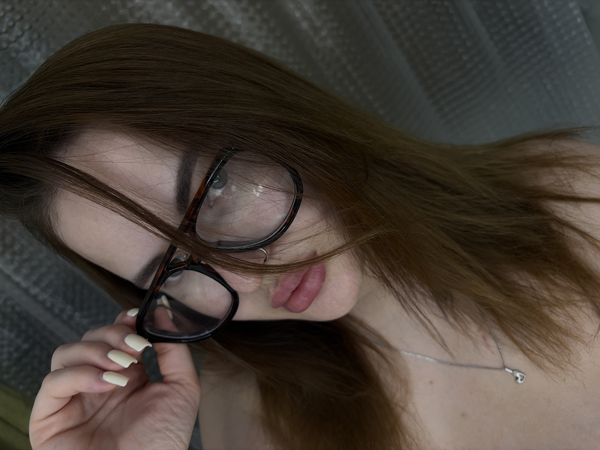 A close-up, tilted portrait of a young woman with brown hair. She is wearing glasses with thick, dark frames. Her left hand is holding the glasses near her face, and her nails are painted with a light color. A silver necklace with a heart-shaped pendant is visible on her neck. The background appears to be a textured, light-colored surface. There is no indication of the location being Krasnodar, Russia.
