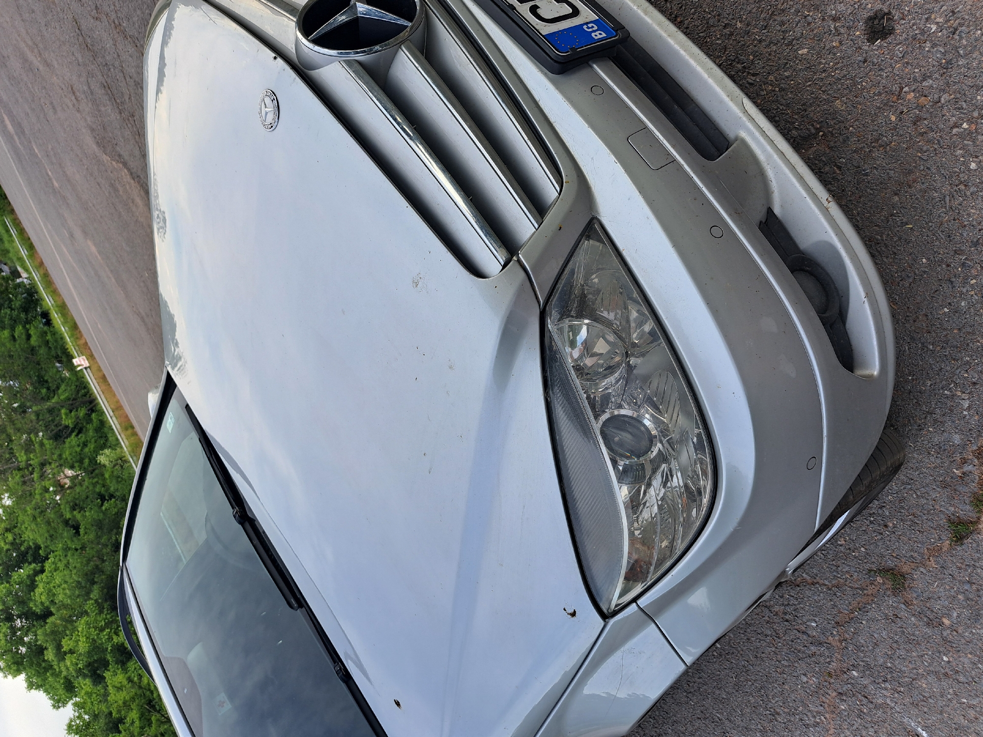 The image is a close-up, low-angle shot of the front right side of a silver Mercedes-Benz sedan parked on a paved road. The car is dusty, with visible specks on the hood and fender. The license plate, which appears to be Bulgarian (indicated by "BG"), is partially visible at the top right, displaying some blue and white markings. The car's grille and headlight are prominently featured. The headlight lens is clear, revealing the internal reflectors and bulbs. The scene appears to be outdoors, with a strip of green trees visible along the left edge of the frame and the asphalt road continuing into the background. The lighting suggests it could be daytime, possibly overcast, given the diffused light. There are no people visible in the frame. The overall impression is a mundane depiction of a parked vehicle.