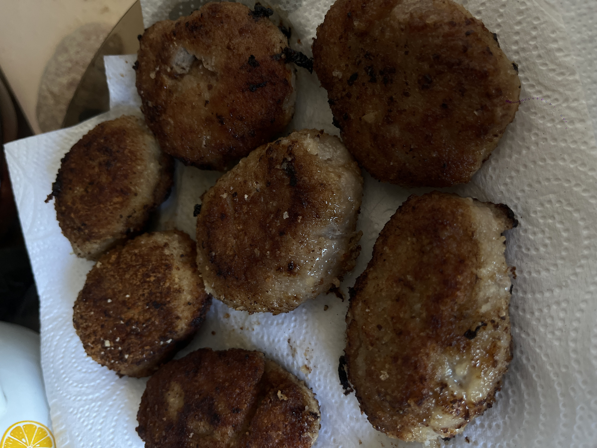 A top-down shot depicts seven cooked, breaded cutlets resting on a white paper towel. The cutlets are irregularly shaped, browned, and appear to be slightly greasy. Some have darker, almost burnt spots. The paper towel is crumpled and shows signs of absorbing oil. Part of a box is visible on the top-left of the image. The bottom-left corner has a partial view of an object featuring an orange citrus fruit design. The location is not possible to be deduced.