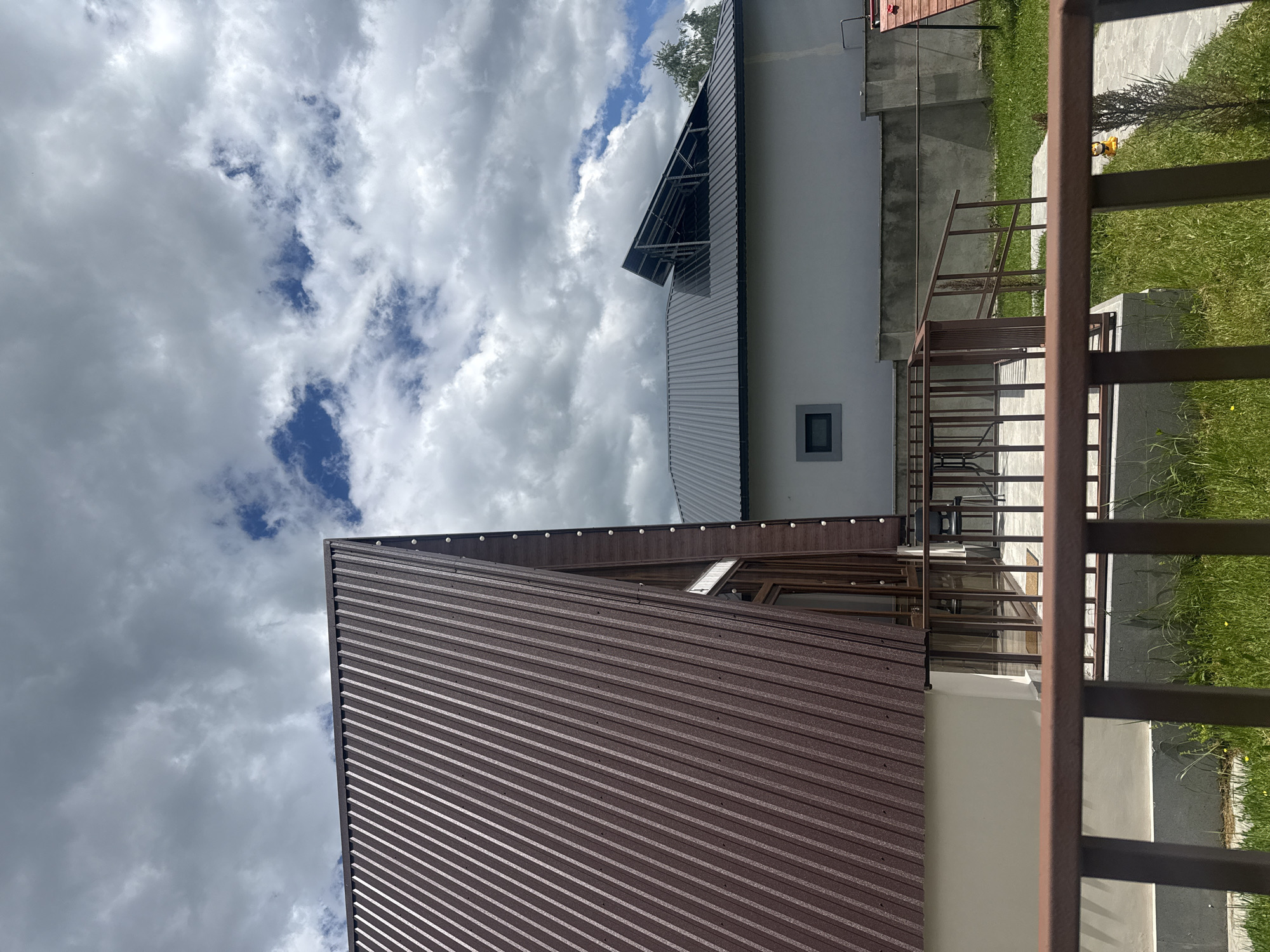 The image depicts a view of two buildings in Stepanavan, Armenia under a cloudy sky. The foreground is dominated by the brown metal roof of a building. Adjacent to it is a white building with a metal roof that has a projecting section and a small, square window. A brown metal fence is partially visible on the right side of the image, obscuring the view of green grass and concrete steps. The cloudy sky occupies the upper portion of the image.
