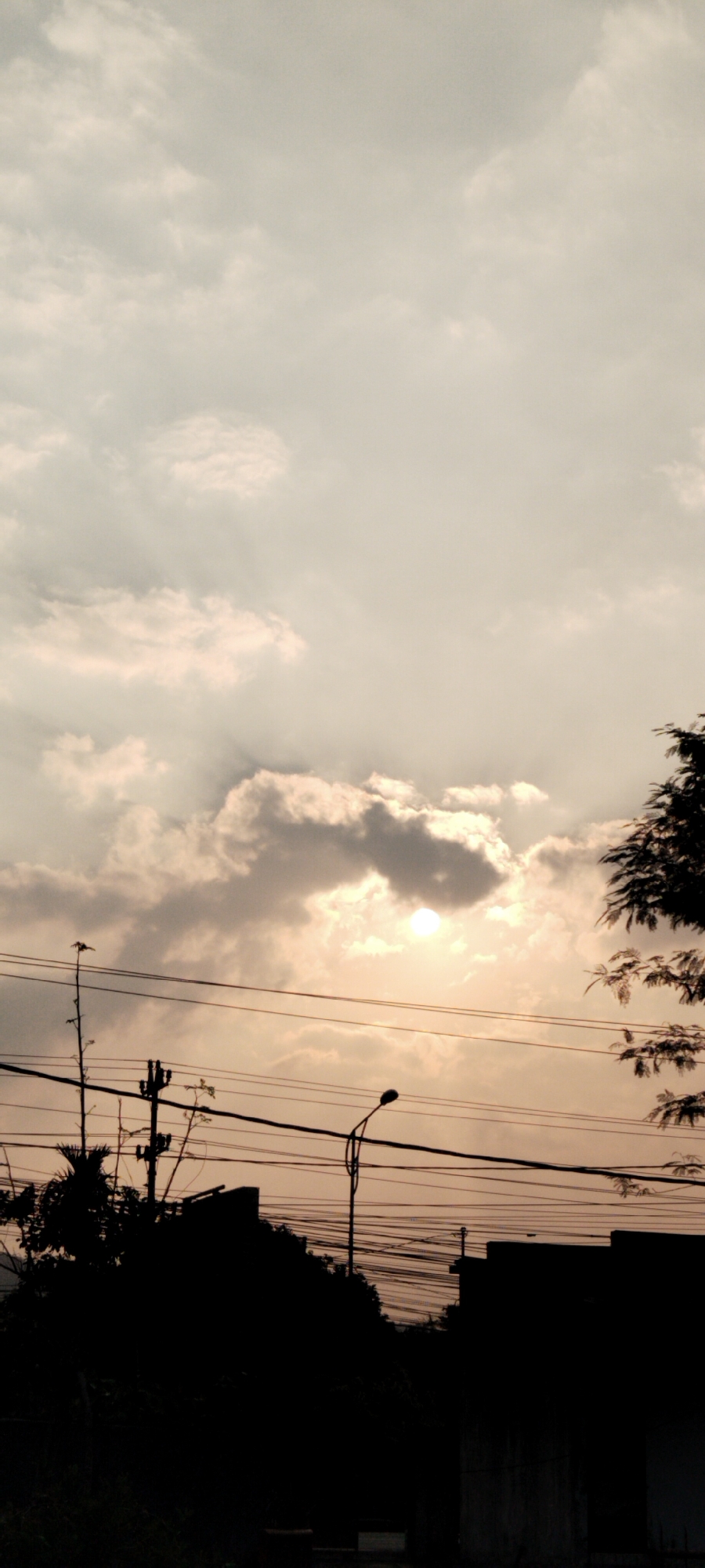 The image depicts a silhouetted landscape against a cloudy sky at dusk in Genuk Barat, Indonesia. The sun is partially obscured by clouds, casting a soft, diffused light. Power lines crisscross the scene horizontally, supported by utility poles. Silhouetted structures, possibly buildings and trees, are visible in the foreground, with dense foliage obscuring some details. A street light stands on the right side, also in silhouette. The overall impression is one of urban twilight, with man-made infrastructure dominating the lower half of the frame and the natural element of the sky above.