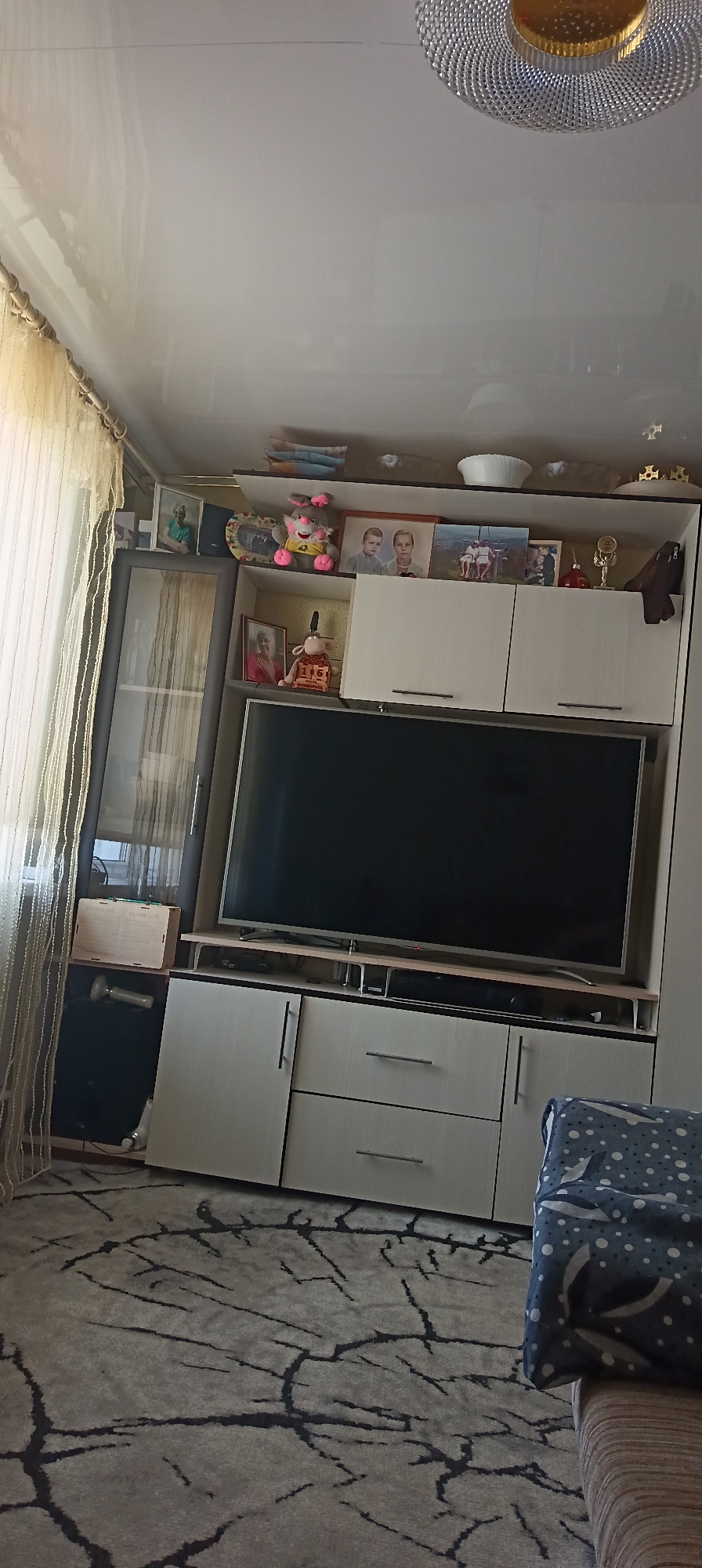 The image depicts an interior living room scene. Dominating the center is a large flat-screen television mounted on a media console. The console has several drawers and cabinets. Above the television, a shelf displays various decorative items, including framed photographs, stuffed animals, and trophies. To the left of the media console is a tall glass-fronted cabinet, also containing displayed items. A beige curtain hangs to the far left of the image, partially obscuring a window. The floor is covered with a patterned rug. The ceiling features a light fixture. The overall impression is of a domestic living space, likely a living room.