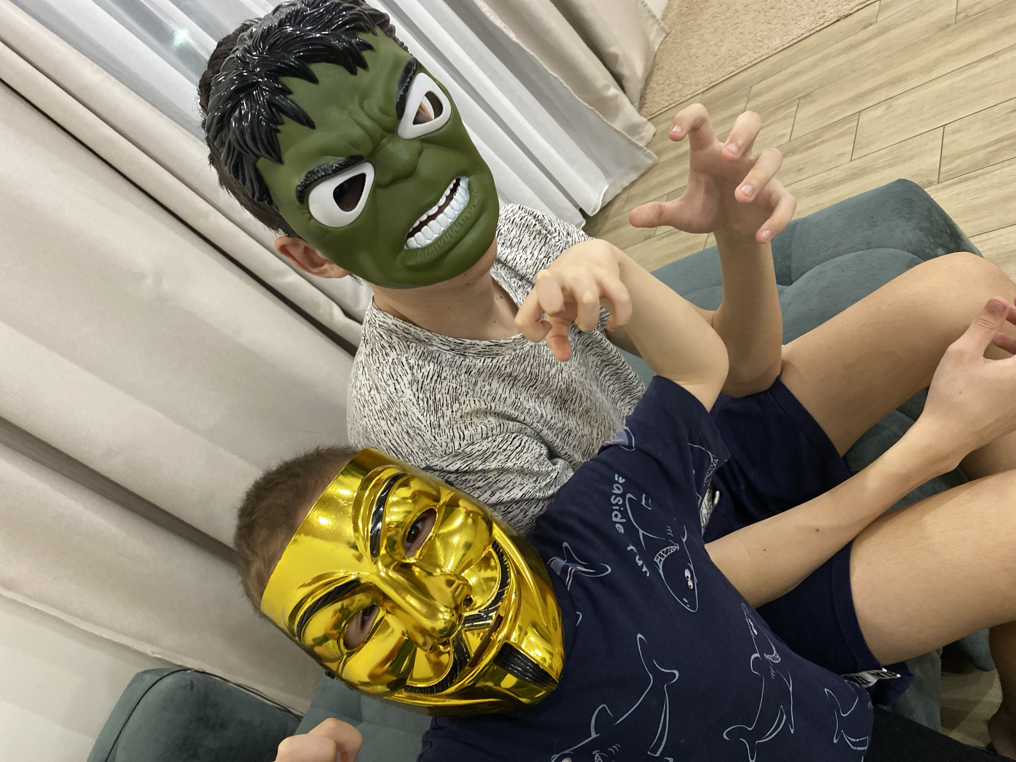 Two children are indoors, both wearing masks. One child wears a green Hulk mask and sits behind the other. The second child lies in front and wears a gold-colored Guy Fawkes mask. The child in the Hulk mask has their hands raised and curled, as if to mimic claws. The location is identified as Chaikovsky, Russia, although that is not evident in the image. The background is a room with a cushioned couch and a curtain.