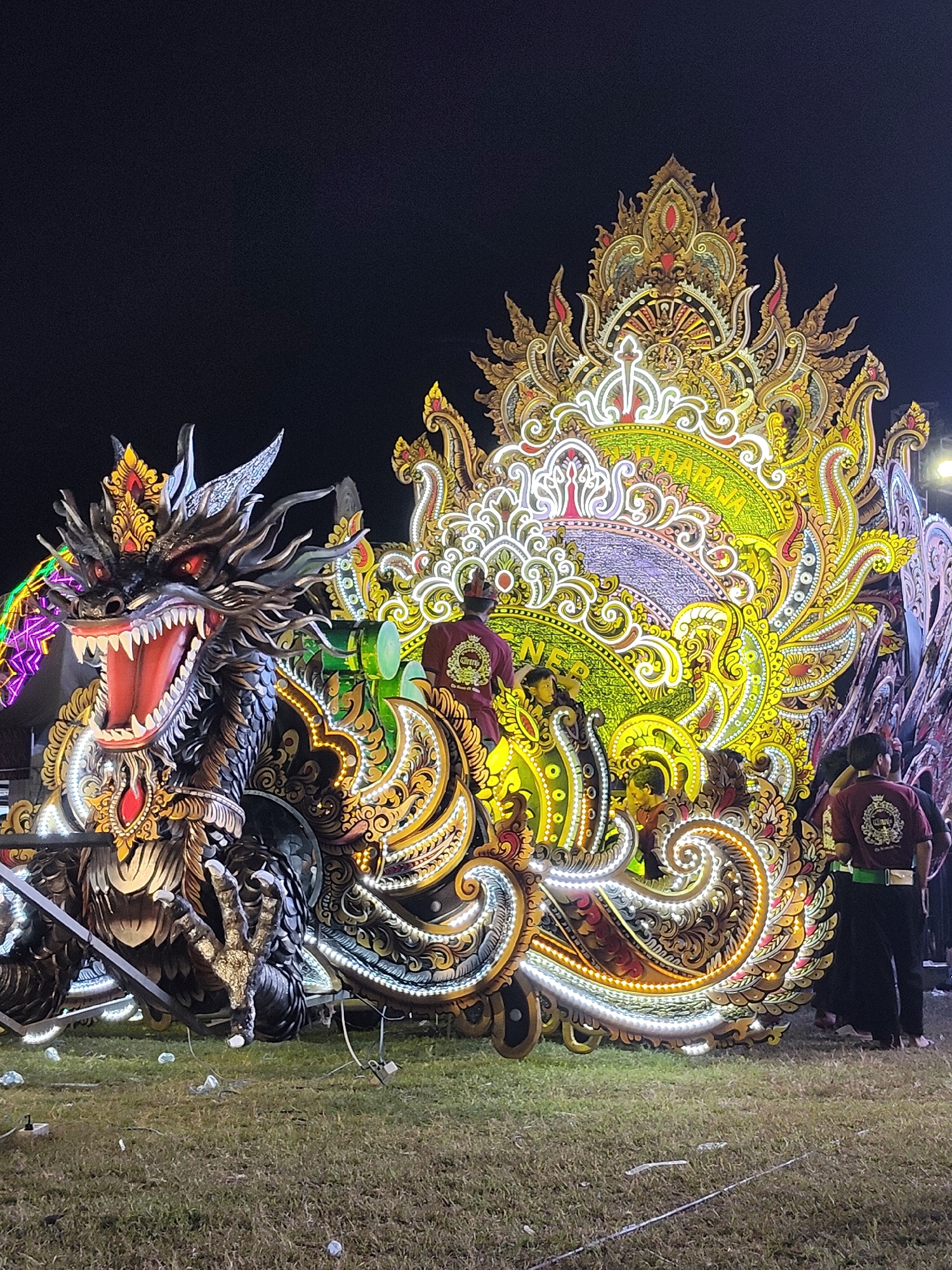 An elaborate, illuminated float is displayed at night in a grassy field in Sumenep, Indonesia. The float features a large, black dragon head with red eyes and an open mouth filled with sharp teeth. The dragon is positioned to the left of a large, ornate, golden and green structure decorated with numerous lights. Several people, dressed in dark pants and maroon shirts with a circular emblem, are visible standing near and on the float. The ground is covered in dry grass with some debris scattered around. The background is dark, indicating it is nighttime.