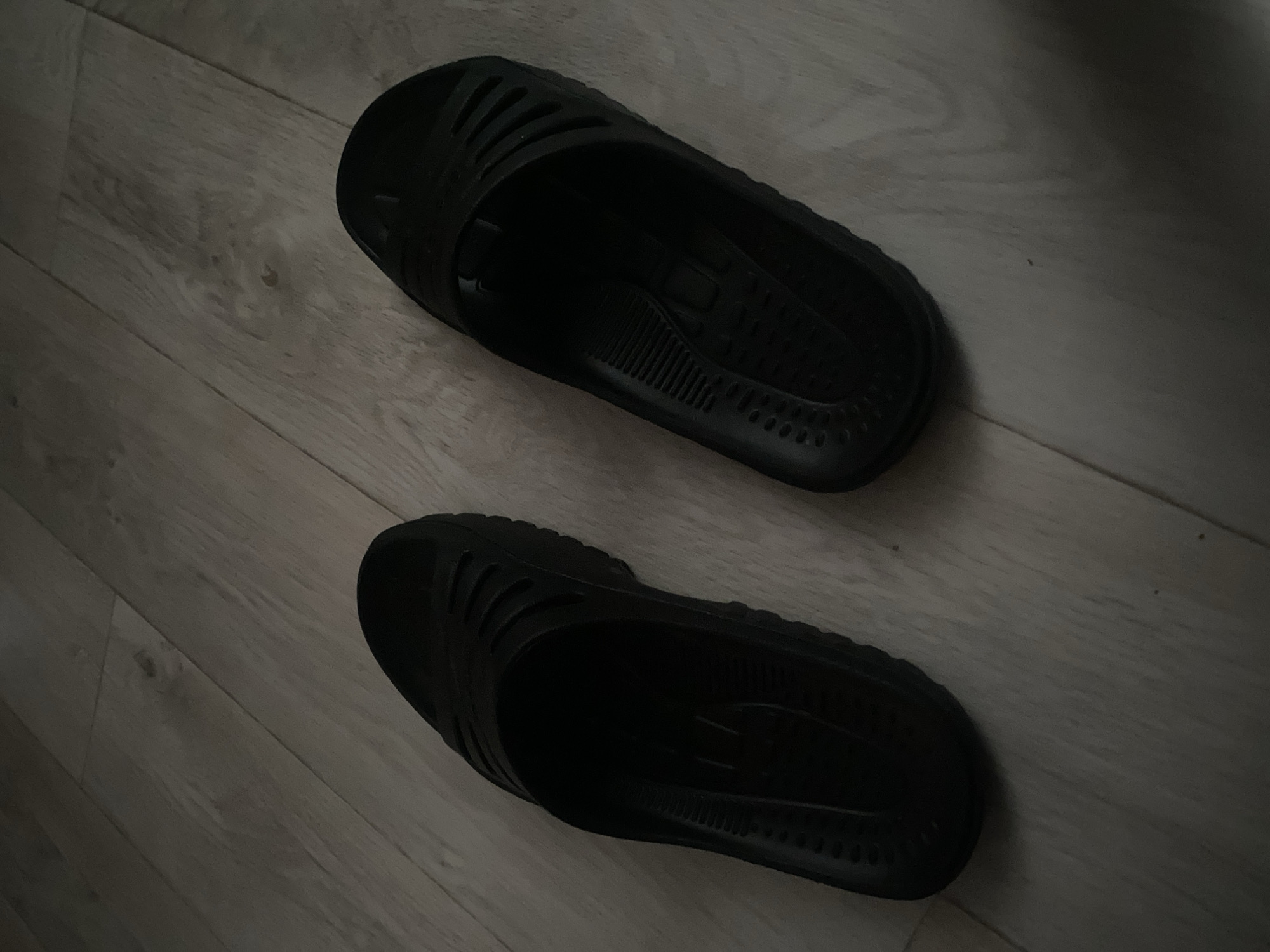 Two black sandals are lying on a light wood-grain floor. The sandals are oriented in slightly different directions, with one shoe angled upwards and the other angled to the left. The floorboards run diagonally in the image. The scene is dimly lit. The image doesn't provide any information about the city of Chaikovsky, Russia.