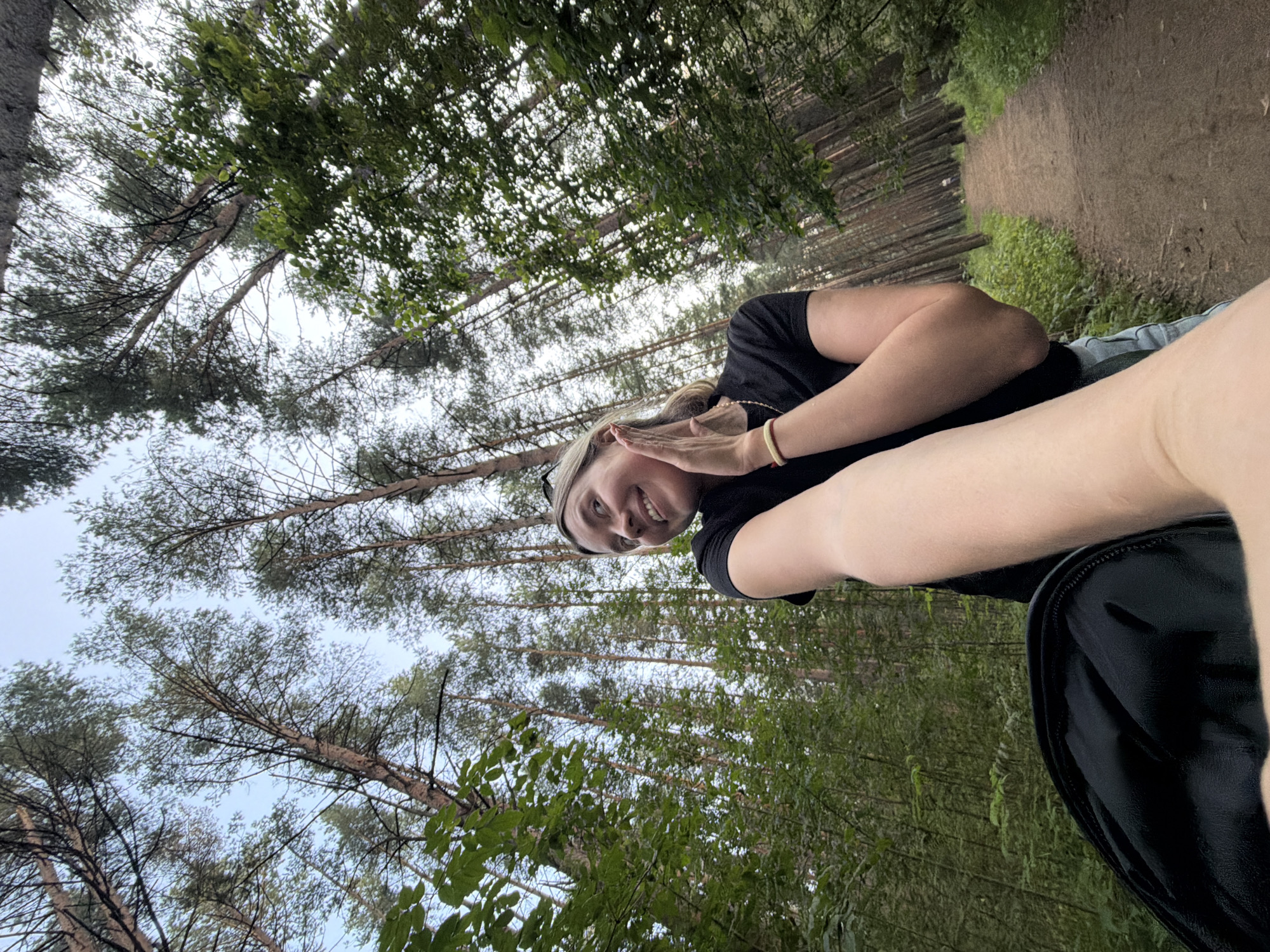 The image depicts a woman in a black t-shirt, wearing light-colored denim shorts, posing for a selfie in a forest. She is smiling and has her left hand gently touching her cheek. Her blonde hair is pulled back. She is wearing a thin bracelet on her right wrist. In the foreground, a black backpack is visible. The background shows a dirt path winding through a dense forest of tall pine trees. The sky is visible through the canopy. The location is a forest trail in Chaikovsky, Russia, characterized by its tall pine trees and a well-trodden path.
