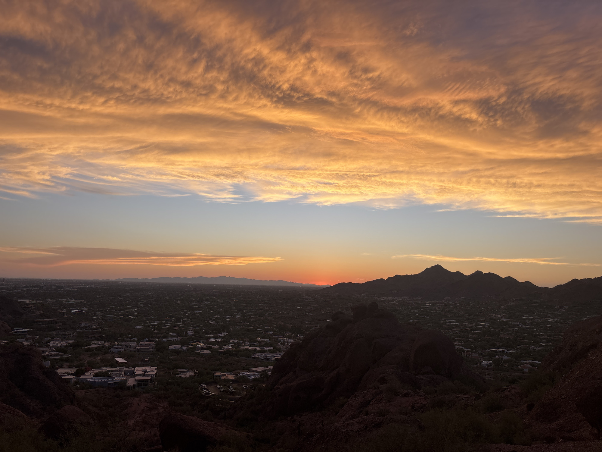 A panoramic, elevated view captures a desert city at sunset. The sky is ablaze with streaks of orange and yellow clouds, forming a dramatic backdrop. In the foreground, rocky, reddish-brown terrain with sparse vegetation suggests a desert environment. Below, a sprawling urban landscape of houses and buildings stretches towards a distant mountain range silhouetted against the horizon. The sun, a bright orange orb, is setting just above the horizon, casting long shadows over the city. The scene appears to be from a vantage point on a hill or mountain overlooking the city, likely in the Phoenix, Arizona area due to the distinctive desert topography and architecture visible. There are no people or active events depicted in the image.