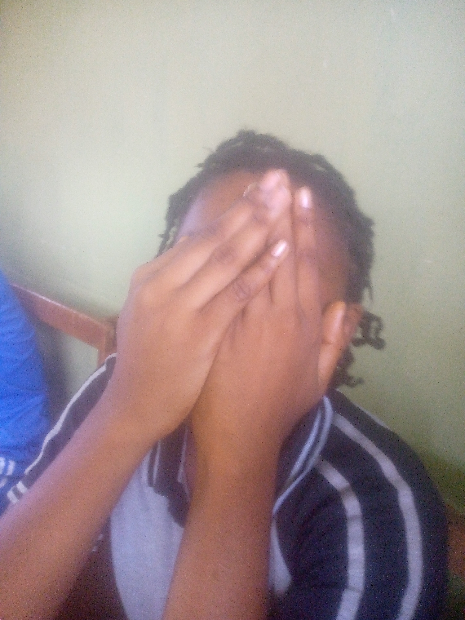 A person with dark skin and dark hair covers their face with their hands. The person is wearing a collared shirt with gray and black vertical stripes. Part of a blue shirt is visible on the left side of the image. The background is a light green wall. The location is Unknown, Unknown.