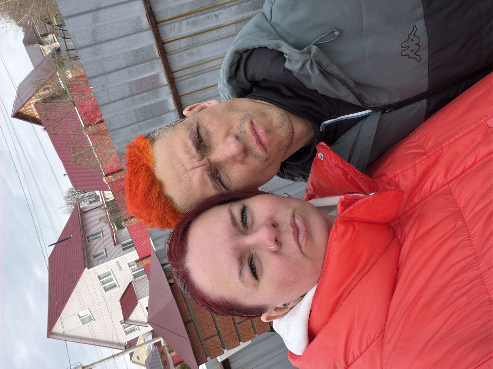 Two people are closely positioned in the foreground of the image. The person on the left has orange hair and is wearing a grey and black hooded jacket with a Kappa logo. The person on the right has reddish-brown hair and is wearing a red padded jacket over a white garment. Both are looking toward the camera with a somewhat neutral expression.
In the background, there are several buildings, including what appears to be a residential structure with a red roof and white siding. The sky is overcast.
Based on available information, it is not possible to determine the precise location in Chaikovsky, Russia.