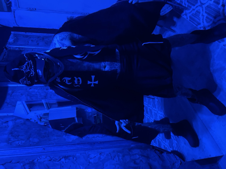 A person stands in a blue-lit room, wearing a black hooded garment with a visible tattoo on their stomach. Their face is partially covered by a mask. The attire features a white cross symbol and lettering, potentially gothic in style. Additional tattoos are visible on the arms and chest. The scene is dominated by blue hues, creating a dark, shadowy atmosphere.