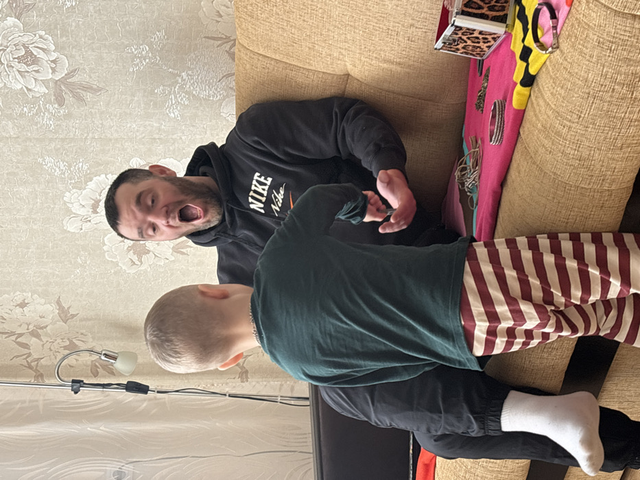 In an interior setting, identified as Pavlovsky, Russia, two individuals are present. An adult male, with short dark hair and a beard, is seated on a beige fabric couch. He wears a black hoodie with "NIKE" printed in white on the front and dark-colored pants. His mouth is wide open, suggesting an exaggerated expression. His left hand is open, palm facing upwards, extended towards the child. A young male child, with light hair, stands facing away from the camera, positioned directly in front of the seated adult. The child wears a dark green long-sleeved shirt, red and white horizontally striped pants, and a silver-colored chain around his neck. His right hand is seen interacting with the adult's outstretched left hand. His left foot, covered by a white sock, is visible.
On the couch between the two individuals and to the right of the child, a pink cloth or blanket covers a section. On this surface, various items are scattered, including multiple strands of beaded or metallic chains, a silver-colored bangle bracelet, and a small leopard-print box with a reflective top. A yellow and black striped fabric item is also partially visible on the couch. To the far left of the image, a metal floor lamp with a white conical shade is standing. The wall behind the subjects is covered with light-colored wallpaper featuring a floral pattern.