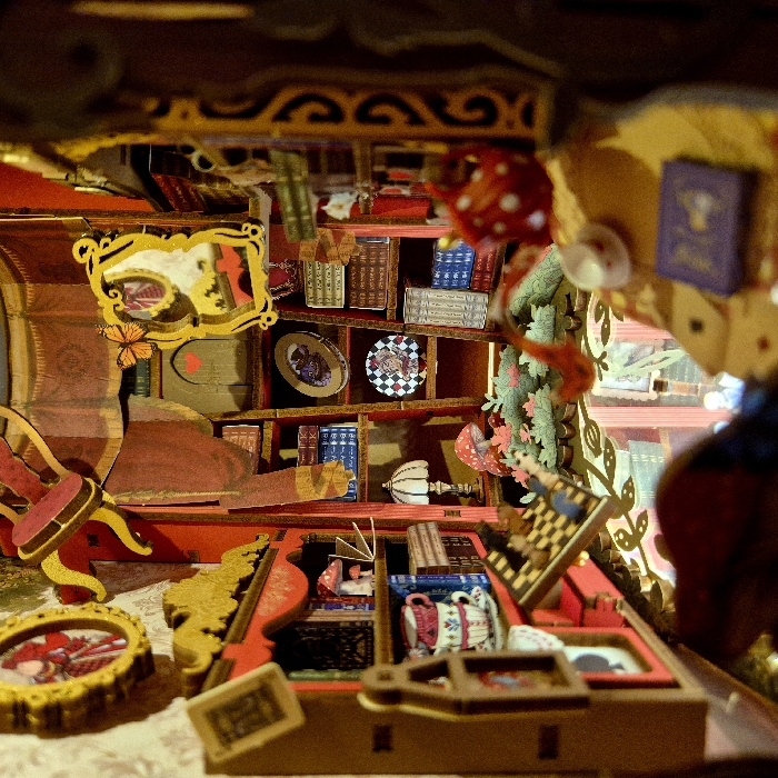 The image displays a highly detailed, miniature diorama or dollhouse scene, crafted with a whimsical, fantastical aesthetic reminiscent of "Alice in Wonderland." The perspective is a downward-looking shot into an intricately designed room, likely a study or a fantastical library.
The scene is rich in warm colors, predominantly reds, golds, browns, and greens, with numerous small, meticulously crafted objects.
**Key elements and objects in the scene include:**
* **Ornate Structures:** The top and upper left sections feature dark, ornate wooden structures with swirling patterns, possibly representing the ceiling or upper architectural details of the miniature house.
* **Bookshelves and Books:** A central feature of the background is a series of miniature wooden bookshelves packed with tiny, colorful books. The book spines are detailed, showing various colors like brown, blue, white, and green, some with gold lettering, though individual titles are too small to decipher clearly.
* **Decorative Wall Elements:**
* On the left wall, a prominent **yellow, ornate picture frame** hangs, reflecting or containing a blurry image of red and gold patterns.
* An **orange butterfly** with black markings is perched on the wall just below this frame.
* **Shelf Decorations:** Among the books on the shelves, several decorative circular plates or objects are visible. One notable item is a **black and white checkered disc or plate**. Another is a darker plate with an indistinct central image. Below these, a white, fluted object, possibly a miniature lamp base or a stylized mushroom, stands on a lower shelf.
* **Whimsical Foliage and Hanging Items:** On the right side of the scene, a vibrant **green vine-like structure** with delicate leaves and hints of pink or red blossoms or berries winds downwards. Suspended from or near this vine are several **red and white polka-dotted mushrooms** (resembling Amanita muscaria) and a small, white **teacup**. Further to the top right, a small **purple book with a gold symbol** on its cover is visible.
* **Furniture and Foreground Details:**
* A miniature **red and gold chair** with a dark base is partially visible on the far left.
* The foreground features a flat surface, likely a desk or table, with an elaborate, multi-tiered **wooden structure** in the center, adorned with gold accents.
* Within this structure and on the surrounding surface, numerous tiny items are arranged:
* Several miniature **teacups**, one prominently displayed with a detailed floral pattern in red, pink, and blue.
* A miniature **chessboard or checkerboard**, tilted open on a stand.
* A small, **ornate gold frame** in the bottom left of the foreground contains a picture of a character in a red dress, possibly Alice.
* Other tiny objects, such as miniature scrolls or decorative pieces, are scattered around.
* **Visible Text:** In the very bottom center foreground, a small rectangular **sign or label** is placed. The text on this sign appears to read "ALICE" in a decorative font.
The overall impression is one of intricate craftsmanship and a fantastical narrative, likely drawing inspiration from Lewis Carroll's "Alice in Wonderland" due to the specific imagery like the checkered pattern, mushrooms, teacups, and the "ALICE" label. The lighting appears to be warm and artificial, highlighting the rich textures and vibrant colors of the miniature world.