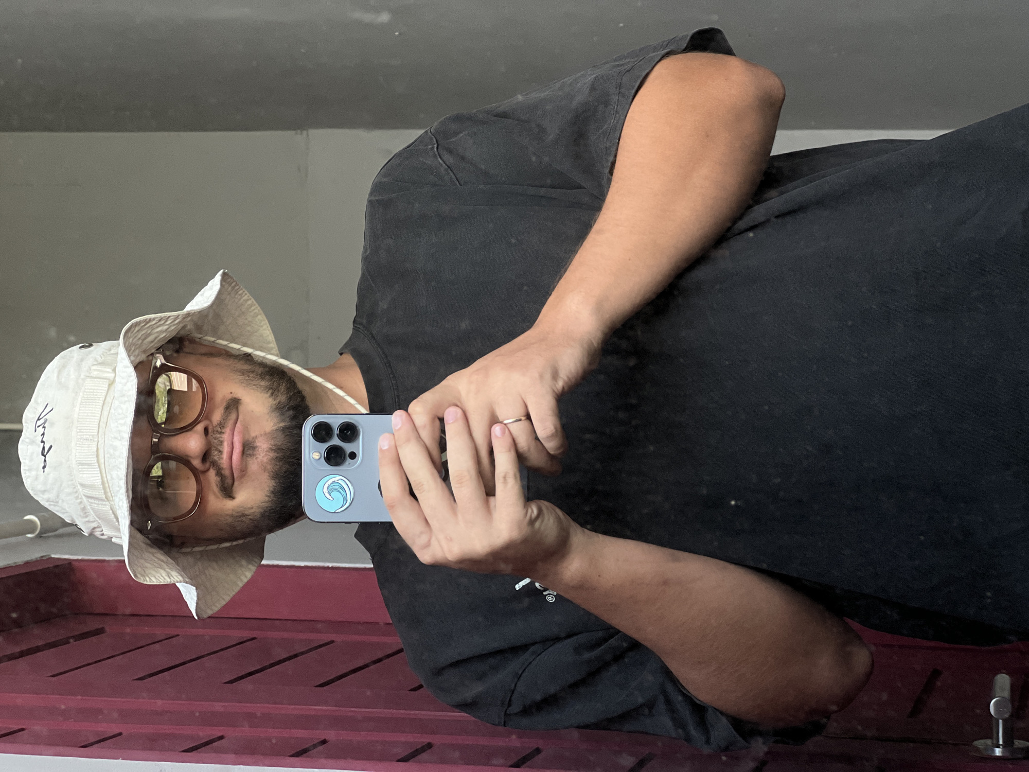 In a medium shot, a person is captured in a selfie from the chest up, holding a smartphone in their hands. The person is wearing a white bucket hat and a black t-shirt, with glasses and a beard. The background features a reddish-brown door on the left and a grey wall on the right, suggesting an outdoor or indoor setting that is not readily identifiable as being in a specific city or location.