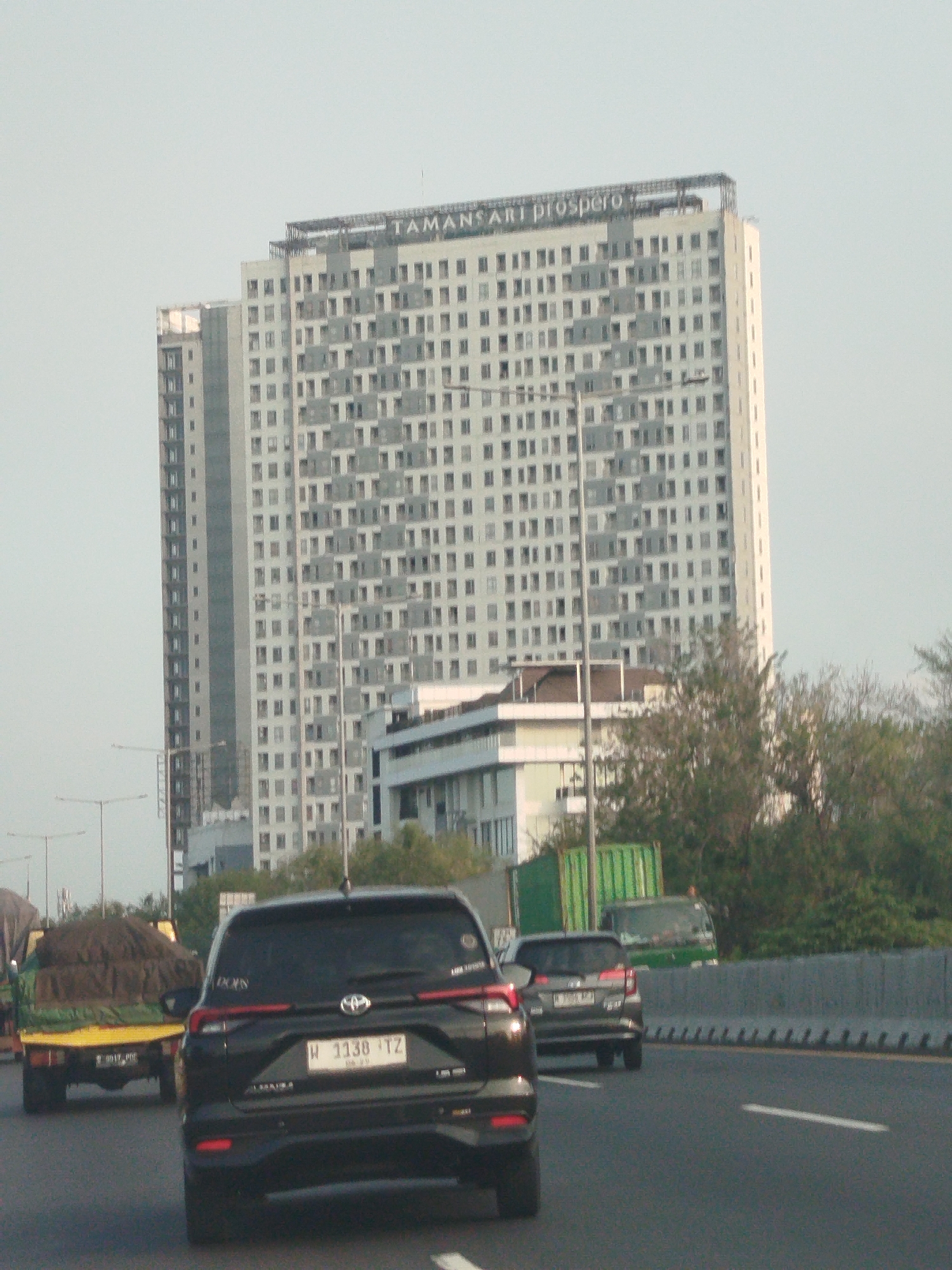The image captures a view from a moving vehicle on a highway in the vicinity of Entalsewu, Indonesia, on a hazy day. The prominent feature is a tall, modern, multi-story building with a sign at the top reading "TAMANSART prospero." The building has a grid-like facade with numerous windows. In the foreground, there is a black SUV with license plate "W 1138 TZ" and another silver SUV. To the left of the SUVs, a yellow truck carrying a large mound of dark material, possibly soil or sand, is visible. The highway is bordered by a concrete barrier. Trees and other vegetation line the side of the road and the area surrounding the building. The sky is overcast and light-colored.