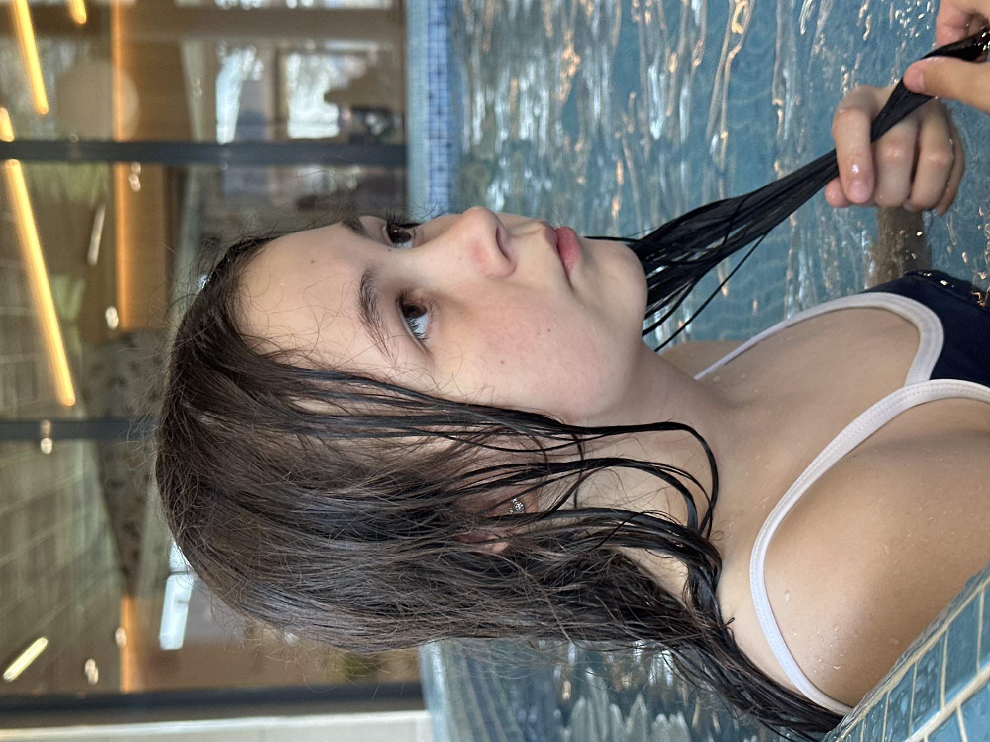 A girl with wet brown hair is in a swimming pool. She is wearing a dark blue swimsuit with white straps. Someone is holding a strand of her hair. The pool is surrounded by blue tiles. A building is visible in the background. The location is unknown and there is no evidence to suggest it is in Votkinsk, Russia.