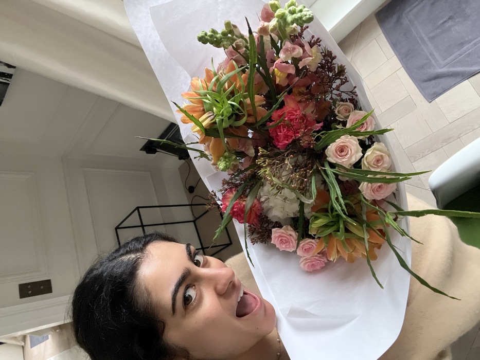 A woman with dark, curly hair is shown in a close-up, tilted selfie, holding a large, vibrant bouquet of flowers. Her face, seen from the forehead down, dominates the lower left and center of the frame. She has wide, open eyes and her mouth is agape, revealing her tongue, suggesting an expression of surprise, excitement, or joy. Her skin tone is fair to medium. She appears to be wearing a light-colored top, possibly beige or cream.
The bouquet of flowers is substantial and elaborately arranged, wrapped in white paper. It features a rich variety of blooms and foliage in shades of soft orange, various pinks, light green, and white. Visible flowers include numerous small, light pink roses, distinctive orange bell-shaped or trumpet-like flowers (possibly Fritillaria) with spiky green foliage emerging from their tops, bright coral-pink ruffled flowers (likely carnations), and clusters of white or cream flowers (possibly hydrangeas). Tall, light green spires (perhaps snapdragons or stock) and darker, reddish-brown foliage add texture and depth to the arrangement.
The setting appears to be indoors, likely a home, with bright natural lighting suggesting daytime. In the background on the left, a white wall with decorative molding is visible. Below the molding, there's a small, rectangular bronze or dark metal plate with two circular openings, possibly an old switch or outlet. Further back on the left, a black, open-frame shelving unit or table is dimly seen. To the right, a light-colored floor, possibly wood or laminate with a herringbone pattern, is visible, along with a grey rectangular mat or towel. A glimpse of a light-colored armchair or sofa, with a white or light grey cushion and a hint of green fabric, is present in the lower right corner. No text is visible in the image.
