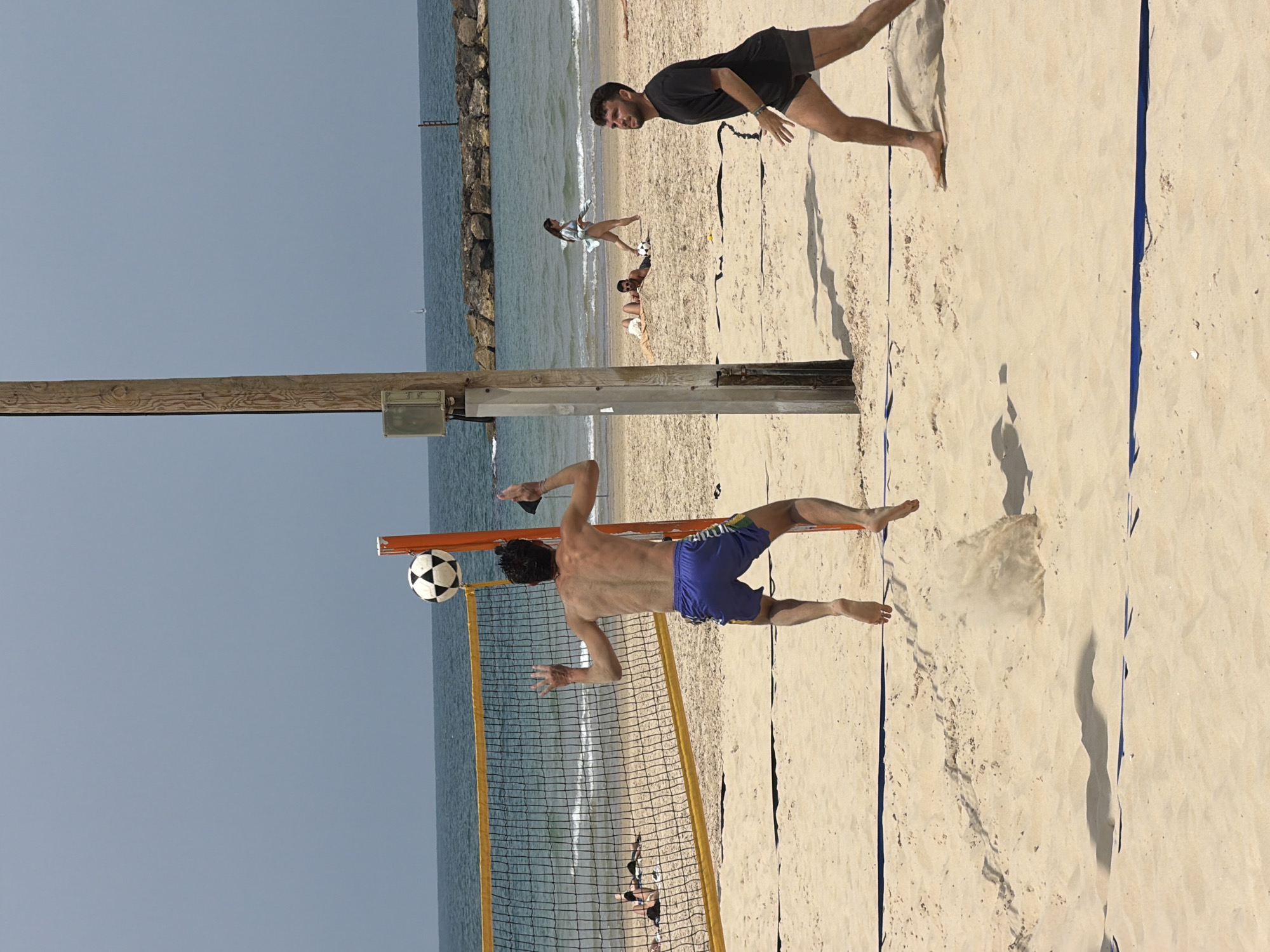 The image captures a beach scene in Tel Aviv, Israel, with a focus on a beach soccer game in progress. A man wearing blue shorts is in mid-air, executing a kick at a soccer ball positioned slightly above a yellow and black net. Another man in black shorts and a black t-shirt is standing further away on the sand. In the background, other people are scattered on the beach, some appear to be sunbathing. The sea and a stone breakwater are visible behind the soccer net, with a clear blue sky above. The sand shows footprints and the shadow of the soccer net and players. A wooden post is positioned near the net.