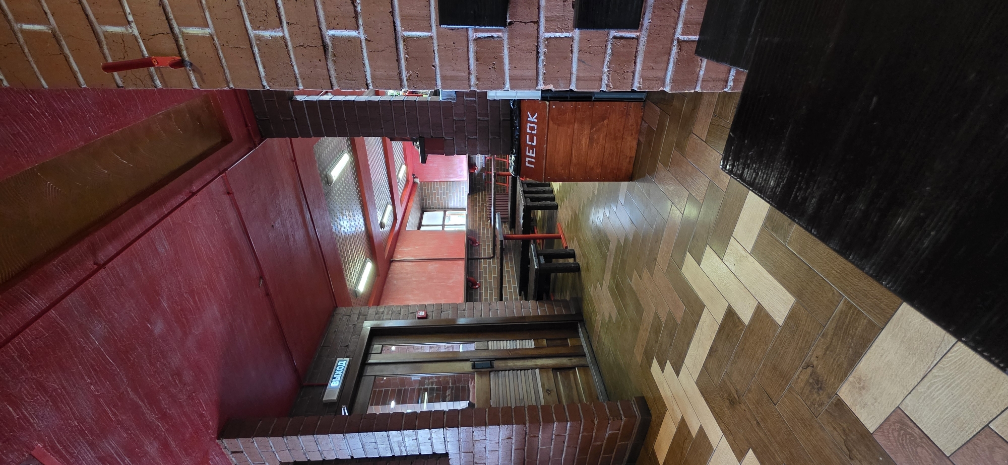 The image depicts the interior of a establishment, likely a cafe or restaurant, in Mezhdurechensk, Russia. The walls are constructed from red brick, with some sections painted red. The ceiling is also painted red, with a textured finish. The floor is tiled in a herringbone pattern using wood-toned tiles of varying shades.
In the foreground, a dark wooden table is visible, with its surface reflecting light. To the left, a doorway with glass panels and wooden frames is present, with a lit "EXIT" sign above it in blue Cyrillic text.
Further into the establishment, rows of dark wooden tables and benches are arranged, separated by brick pillars. Fluorescent lights are mounted on a metallic ceiling in the background, illuminating the dining area. A wooden bin with the Cyrillic word "ПЕСОК" (meaning "sand") printed on it in white is positioned near a brick pillar. The overall ambiance suggests a casual, perhaps industrial-themed dining space. No people are visible in the image.