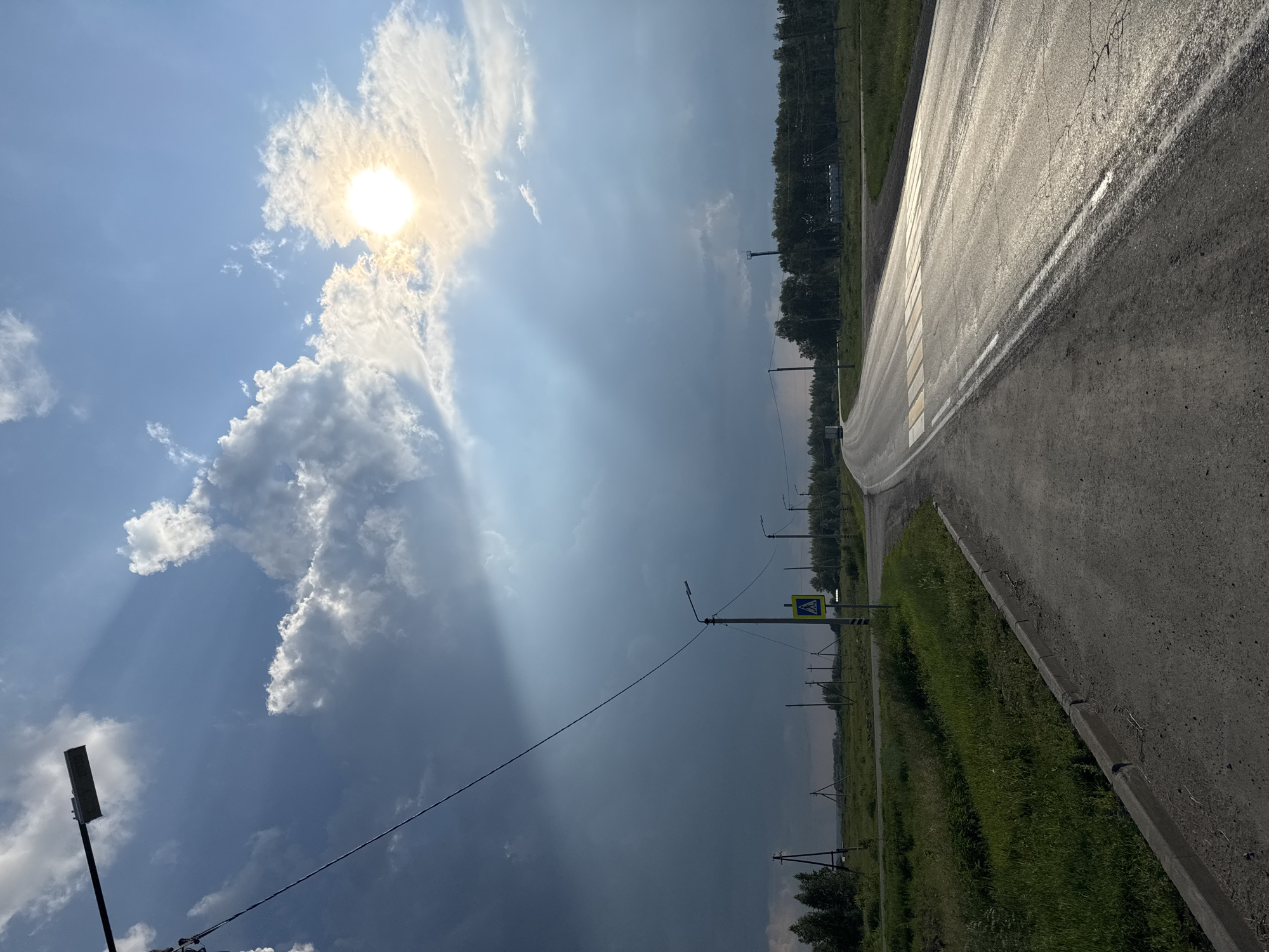 The image depicts a roadside scene in Архангельское, Russia. A paved road curves to the right, with a grassy shoulder and a line of trees in the distance. Overhead, the sky is a mix of blue and grey clouds, with the sun partially obscured by a large cloud. Sunbeams pierce through the clouds, creating dramatic lighting effects. Several utility poles with wires are visible along the side of the road, and one pole has a pedestrian crossing sign. The road surface appears wet, suggesting recent rain. The overall impression is one of a dramatic sky over a rural or semi-rural landscape. The time of day appears to be late afternoon or early evening, given the angle of the sun and the play of light and shadow.