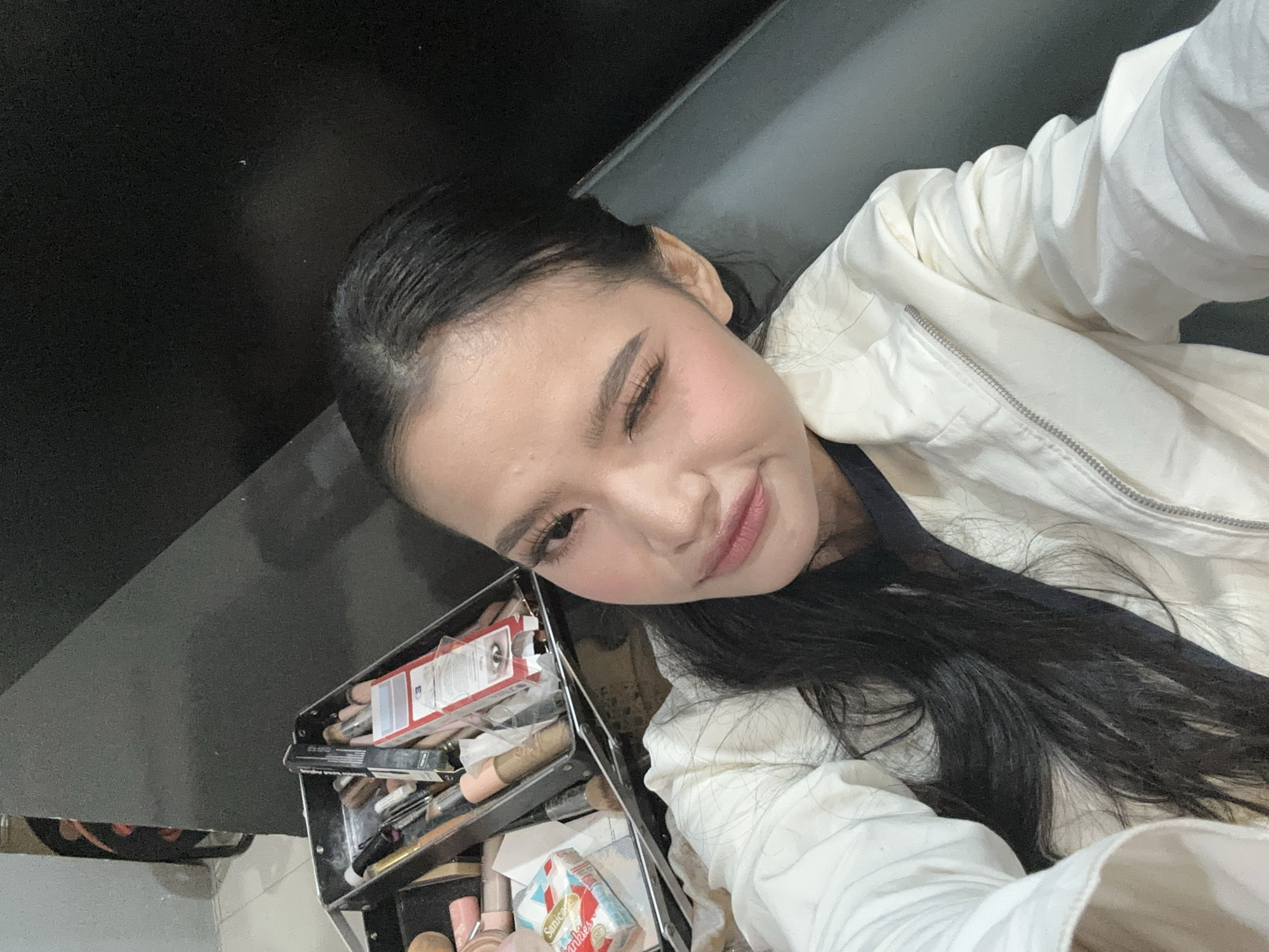 The image is a close-up selfie of a young woman with dark hair, who is winking. She is wearing a white jacket over a dark top. To her left, a black makeup case is visible, filled with various makeup products, brushes, and what appears to be a pack of tissues. The background is dark and out of focus, suggesting an indoor setting, possibly a dressing room or a makeup studio. The presence of the makeup case indicates a context related to beauty or performance. There is no discernible information in the image that would place the location in Davao City, Philippines.