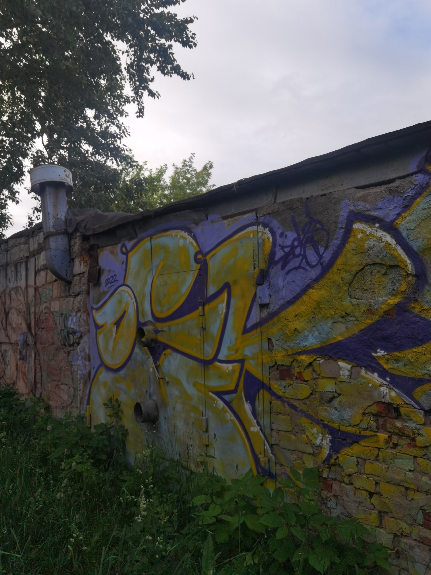 The image shows a section of a weathered brick wall covered in colorful graffiti, located in Chaikovsky, Russia. The graffiti features abstract shapes and lettering in yellow and purple against a light blue and grey background. A metal pipe, likely a vent or drainpipe, protrudes from the wall, appearing rusted and aged. The wall itself is constructed of light-colored bricks, some of which are chipped or missing, revealing older brickwork underneath. A dark, corrugated material covers the top edge of the wall. Overgrown green grass and foliage are visible at the bottom of the frame, suggesting an outdoor, possibly neglected, urban environment. A large tree with dark green leaves partially obscures the sky in the upper left corner. The sky is overcast with grey clouds. The graffiti includes the year "2022" written in purple.