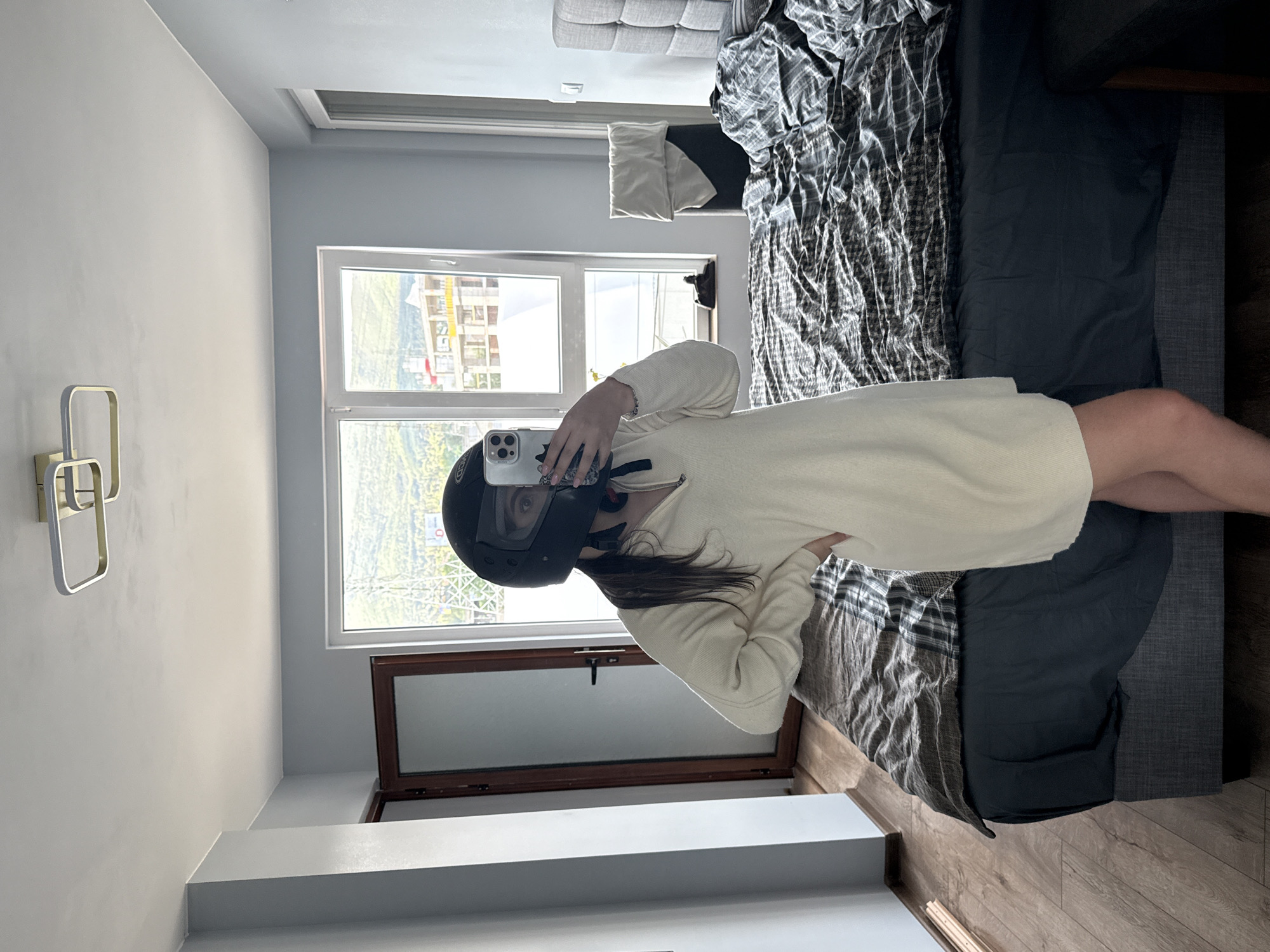 A person is taking a photo of themselves with a smartphone while wearing a black helmet. They stand next to a bed in a room with a window overlooking a building under construction. The photo is taken in Sofia, Bulgaria, based on the architectural style of the buildings visible through the window. The room is well-lit, with a light fixture on the wall. The person wears a light-colored garment. The bed is covered with a grey and white checkered blanket and a dark blue comforter. A shelf is visible below the window. The floor is wooden.