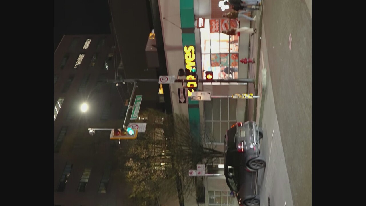 A dark, grainy, night-time, street-level shot captures a bustling urban scene. A dark-colored SUV is parked in the foreground. In the middle ground, a Popeyes Louisiana Kitchen restaurant is prominently lit, with its distinctive orange signage. Several pedestrians are visible outside the restaurant, and traffic lights and street signs indicate an intersection. A streetlamp illuminates the area. The overall impression is that of a typical commercial district at night. The image likely depicts a location in Vancouver, Canada, given the "Granville St" street sign visible in the upper portion of the frame.