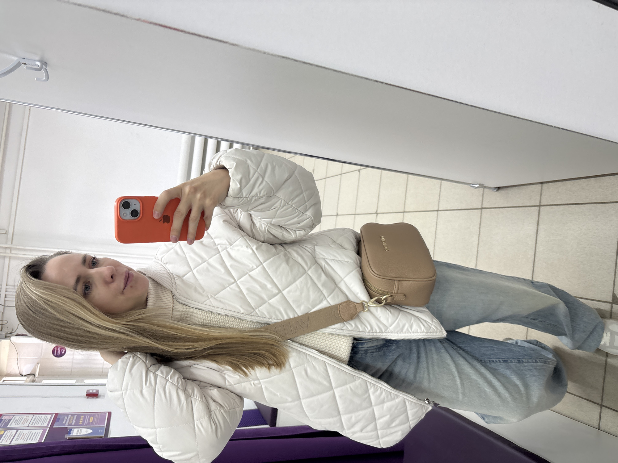 A woman is taking a selfie in a brightly lit indoor space. She is holding an orange smartphone. She's wearing a white quilted jacket over a white turtleneck sweater, and blue jeans. She has a light brown purse slung across her body. She is standing in what appears to be a changing room or waiting area, indicated by the white walls and a purple bench. Based on available information, it's probable that the location is in the city of Chaikovsky, Russia. Details like the signs on the wall suggest a public space such as a hospital. The floor is tiled.
