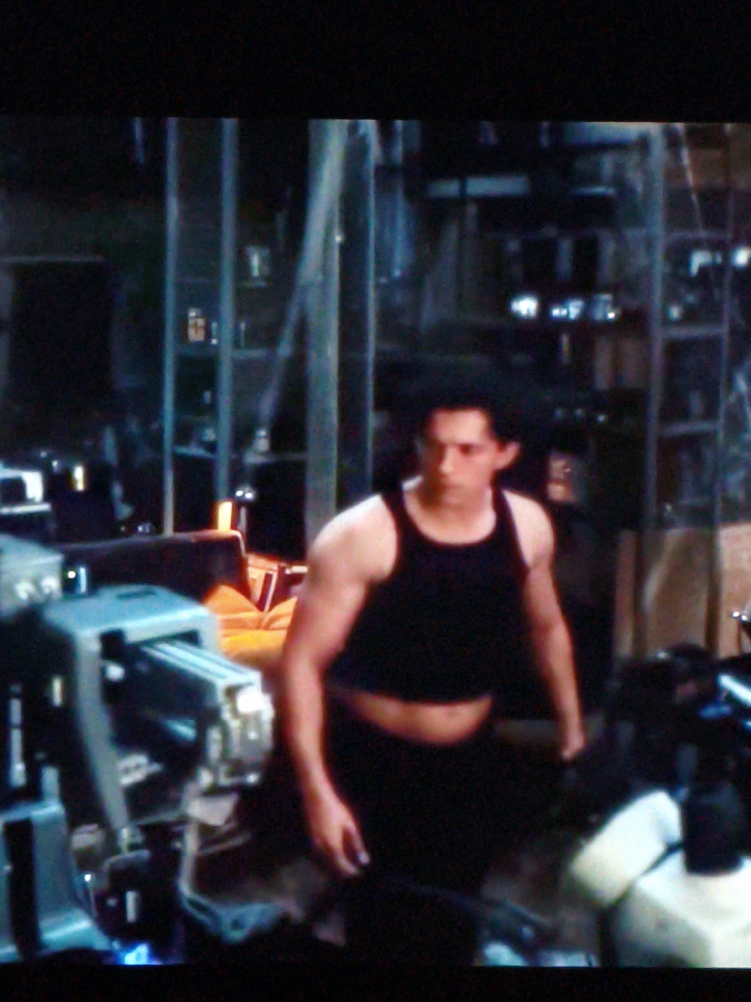The image shows a young man in a black tank top and black pants. He is standing in what appears to be a workshop or laboratory, with shelves filled with various items in the background. To his left, there is a large, complex piece of machinery, possibly industrial equipment. The lighting is dim, with some blue light emanating from the machinery, suggesting it might be in operation or have some illuminated components. The man's expression is neutral, and his posture is relaxed. The scene seems to depict a moment of quiet or pause within a busy environment. There is no visible text in the image. The setting does not provide specific cues that would place it definitively in Vladivostok, Russia, beyond the general context provided.