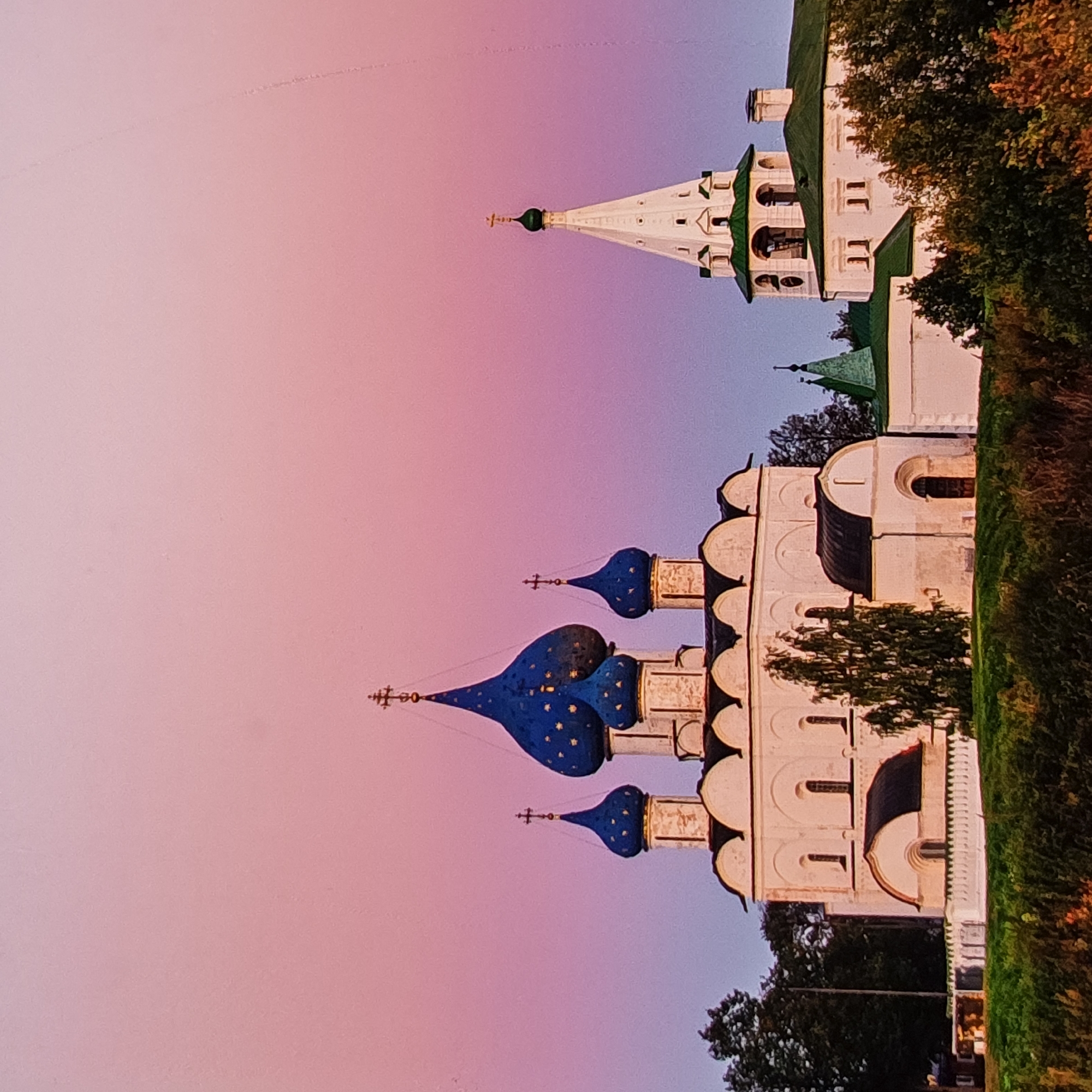 The image depicts a cluster of white churches with blue domes and a tall, white bell tower with a green roof. The architecture suggests a Russian Orthodox religious site. The sky is a gradient of soft pink and purple, indicating a sunrise or sunset. The foreground is filled with lush green foliage and trees. The buildings are situated on a gentle rise, with a white fence in front. The overall impression is one of serene beauty and historical significance. This scene is likely the Suzdal Kremlin in Vladimir Oblast, Russia, a well-known architectural ensemble. There are no people or animals visible in the image. The lighting and colors suggest a calm, picturesque moment.