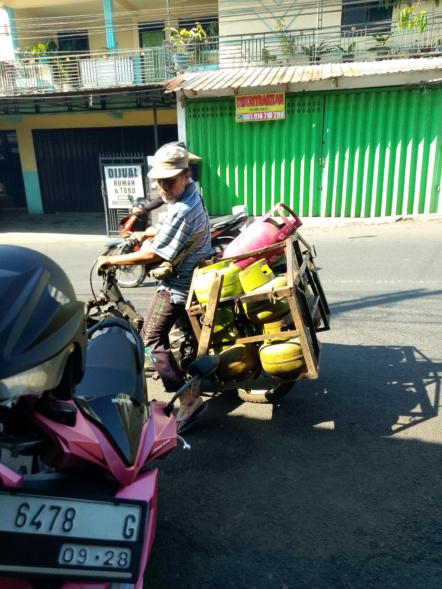 In the city of Gebang, Indonesia, a man wearing a striped shirt, cap, and dark pants is riding a motorcycle. The motorcycle has a metal rack attached to the back, filled with several propane gas cylinders. Some cylinders are red, and others are yellow. The man is partially obscured by the gas cylinders and the rack. The background features a building with a green shuttered entrance, a sign that reads "DIKONTRAKKAN HUBUNGI: 081 914 710 299", and another sign that says "DIJUAL RUMAH & TOKO". Another motorcycle, partially visible, is in front of the man. The scene appears to be on a street, with a paved surface and shadows cast by the sun. The license plate on the front motorcycle reads "6478 G 09-28".