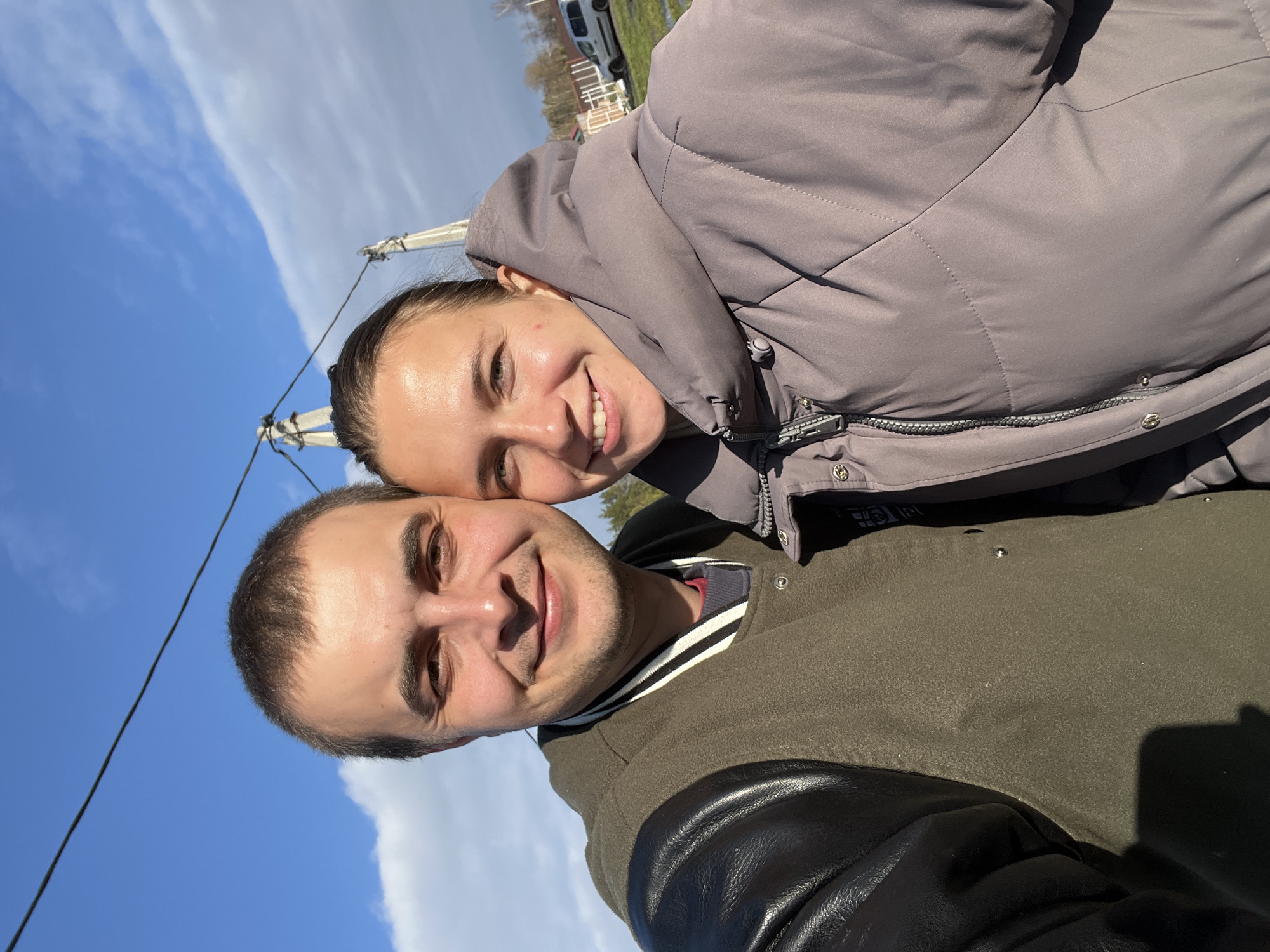 A brightly lit outdoor selfie captures two young adults, a man and a woman, smiling under a clear blue sky with scattered white clouds. The image is oriented sideways but depicts an upright scene.
In the foreground, the man smiles directly at the camera. He wears a dark olive green jacket featuring black leather sleeves and a striped collar (black and white). His face is well-lit by natural sunlight. Slightly behind him and to his left, the woman rests her head close to his, also smiling warmly at the camera. She is dressed in a light grey or lavender puffer jacket.
The background reveals a expansive blue sky, interrupted by a long, thin band of white clouds and several power lines or wires. Distant, blurred elements in the far background suggest an open, possibly rural or semi-rural, outdoor environment, including a faint white building or vehicle. The bright sunlight indicates it is daytime with pleasant, clear weather. Both individuals convey emotions of happiness and contentment. No legible text is visible in the image. The location context for this scene is Razdolye, Russia.