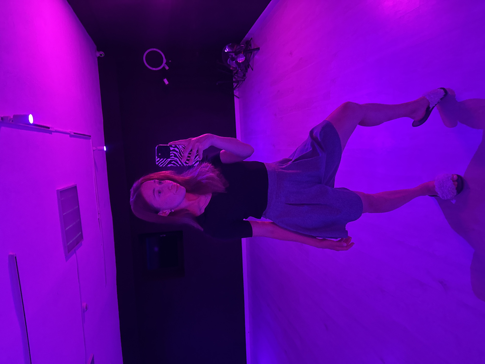 A woman is standing in a room with purple lighting. The walls and ceiling are different colors. The woman is holding a phone with a black-and-white zebra pattern on the case. The background includes a white wall, black walls, and wooden floors. A security camera is visible on the wall. The location is Chaikovsky, Russia.