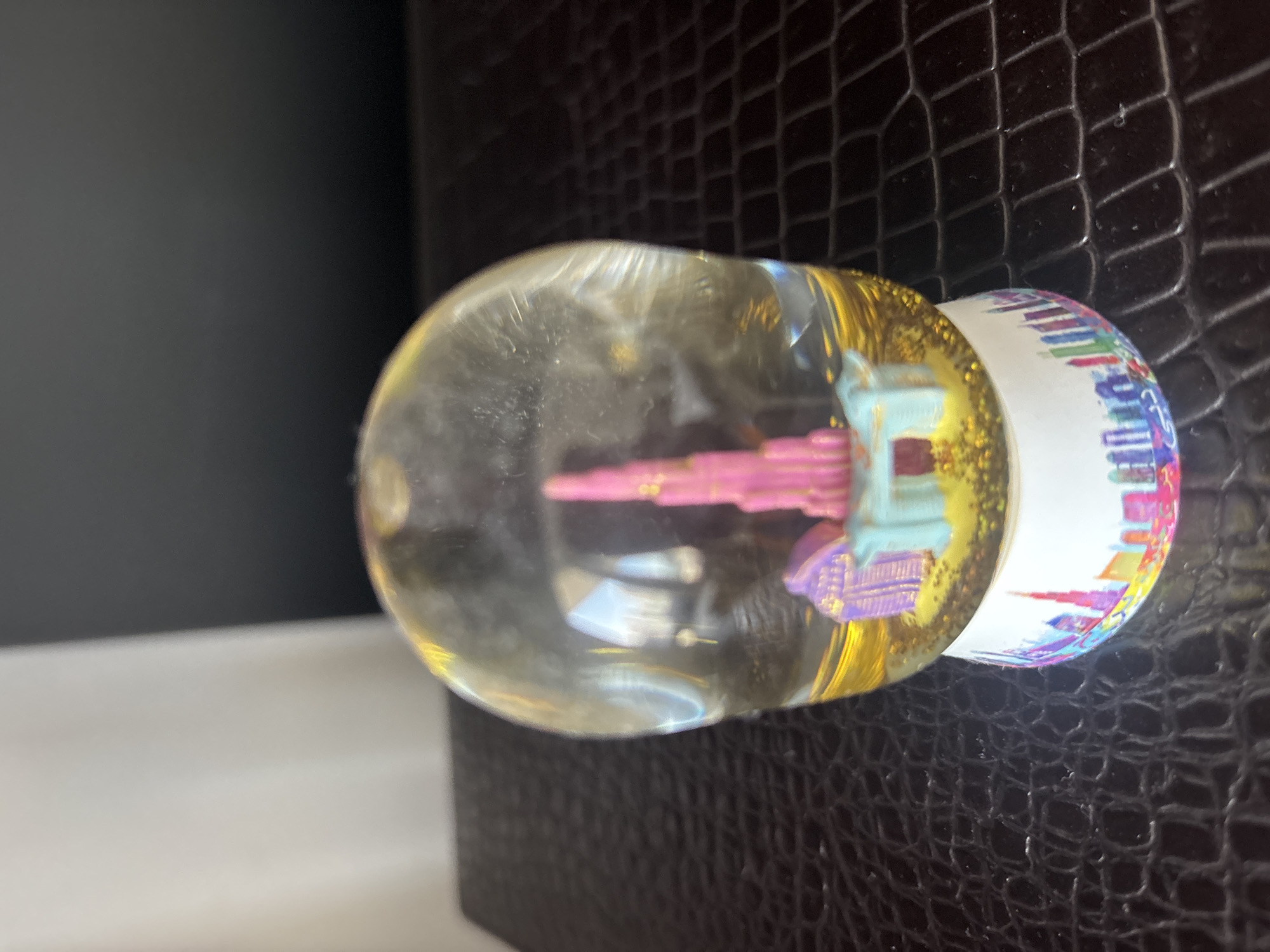 A close-up, eye-level shot shows a souvenir snow globe featuring a miniature representation of the Burj Khalifa, Dubai's iconic skyscraper. The snow globe contains a pink, tiered replica of the Burj Khalifa, surrounded by blue and purple buildings, all suspended in a clear liquid with gold glitter. The base of the snow globe is decorated with a colorful cityscape, also depicting Dubai's skyline. The snow globe rests on a dark, textured surface, possibly a table with a crocodile-skin pattern. The background is softly blurred, with a hint of white fabric on the left and a dark, solid area on the right. The lighting creates a subtle glow around the snow globe.
