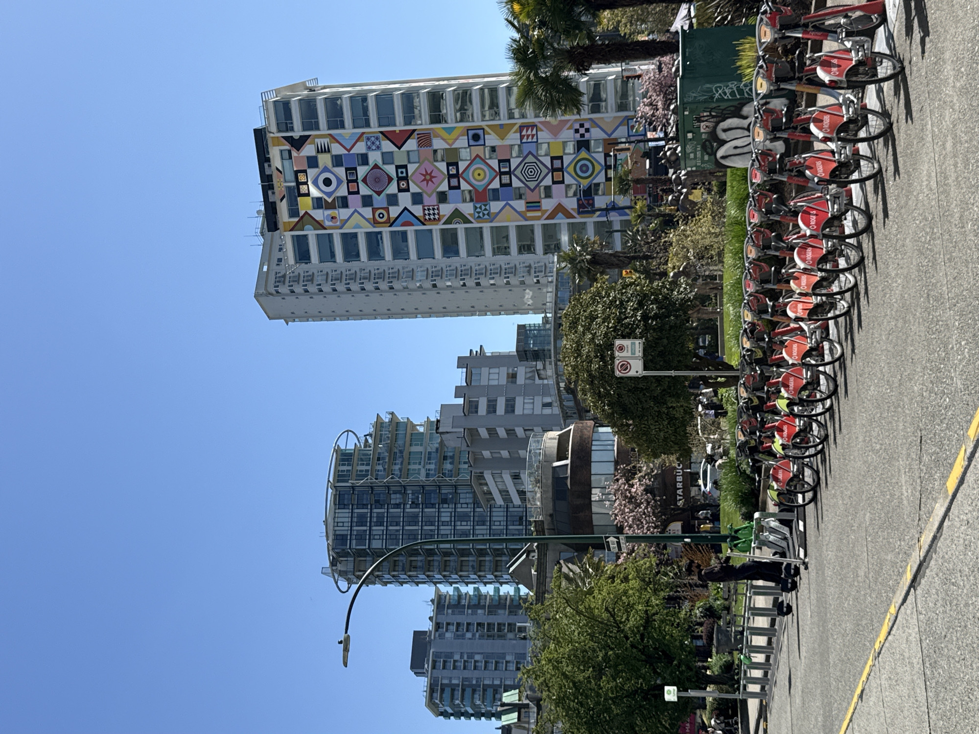 The image depicts a street scene in Vancouver, Canada. In the foreground, a row of red and white bikes are parked along the sidewalk. Behind the bikes is a row of trees and shrubs, followed by a series of tall buildings. One of the buildings is particularly distinctive, with a colorful geometric pattern on its facade. The sky is clear and blue. There is a person walking next to the bikes.