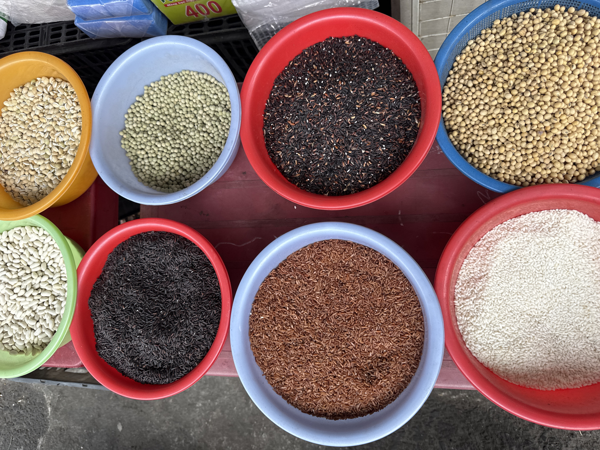 A top-down shot displays nine bowls of different grains and legumes, likely for sale. The bowls are arranged in a grid-like pattern on a red surface. The bowls vary in color (red, blue, yellow, green, light blue) and size, and each contains a different type of grain or legume. Grains include various colored rice (white, brown, dark purple), and legumes include various types of beans (brown, white, light green, and yellow). The image was taken in the city of Đà Nẵng, Vietnam.