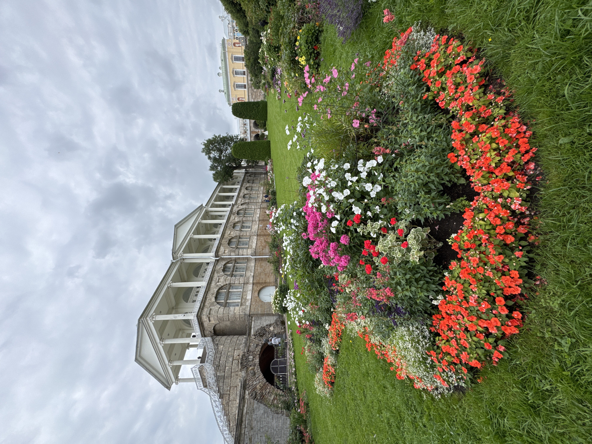 This is a medium-shot, outdoor, eye-level, vertical photograph taken on a cloudy day in the city of Pushkin, Russia, at Catherine Palace's landscape garden. In the foreground, a vibrant, multi-colored flower bed forms a curved border of red, pink, white, and purple flowers with green foliage, set against a backdrop of a meticulously manicured green lawn. Behind the flower bed and to the left, a large, neoclassical stone building with a long, colonnaded white portico and a grand staircase ascends to the upper level. The lower section of this building features an arched stone entryway with a metal gate, behind which a person in a light-colored outfit is walking. To the right of the main building and further in the distance, another yellow building with green rooftops is visible, along with two tall, conical trees. The sky is overcast with gray clouds, contributing to a subdued, diffused light.