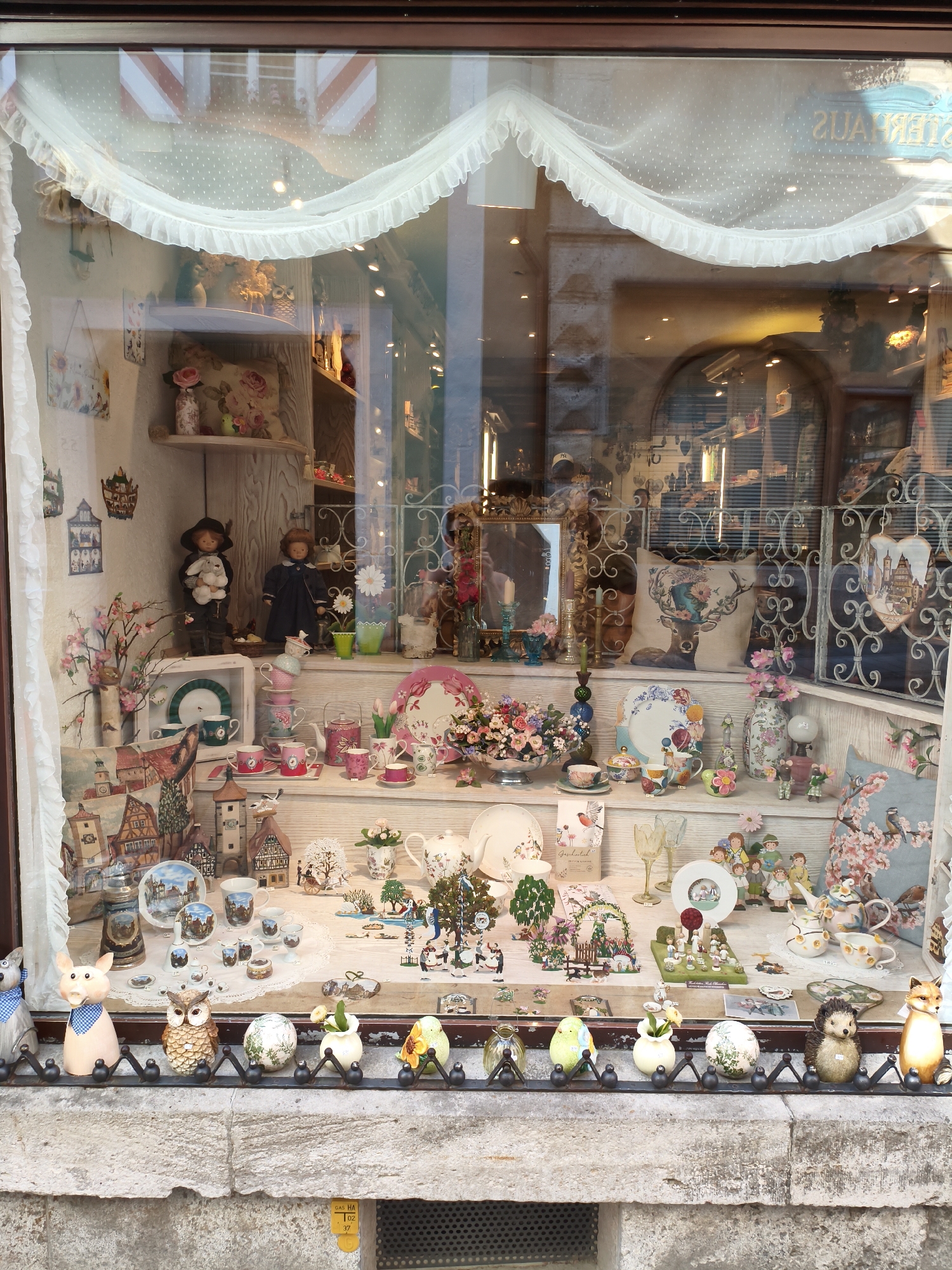 The image shows a display window of a shop in Rothenburg ob der Tauber, Germany. The window is filled with a collection of decorative items, including porcelain figurines, teacups, plates, vases, and miniature houses. The items are arranged on shelves and in the foreground, creating a whimsical and charming display. The window also features decorative white lace curtains. In the background, reflections of the street and buildings can be seen through the glass. The overall impression is one of a quaint and old-fashioned shop.