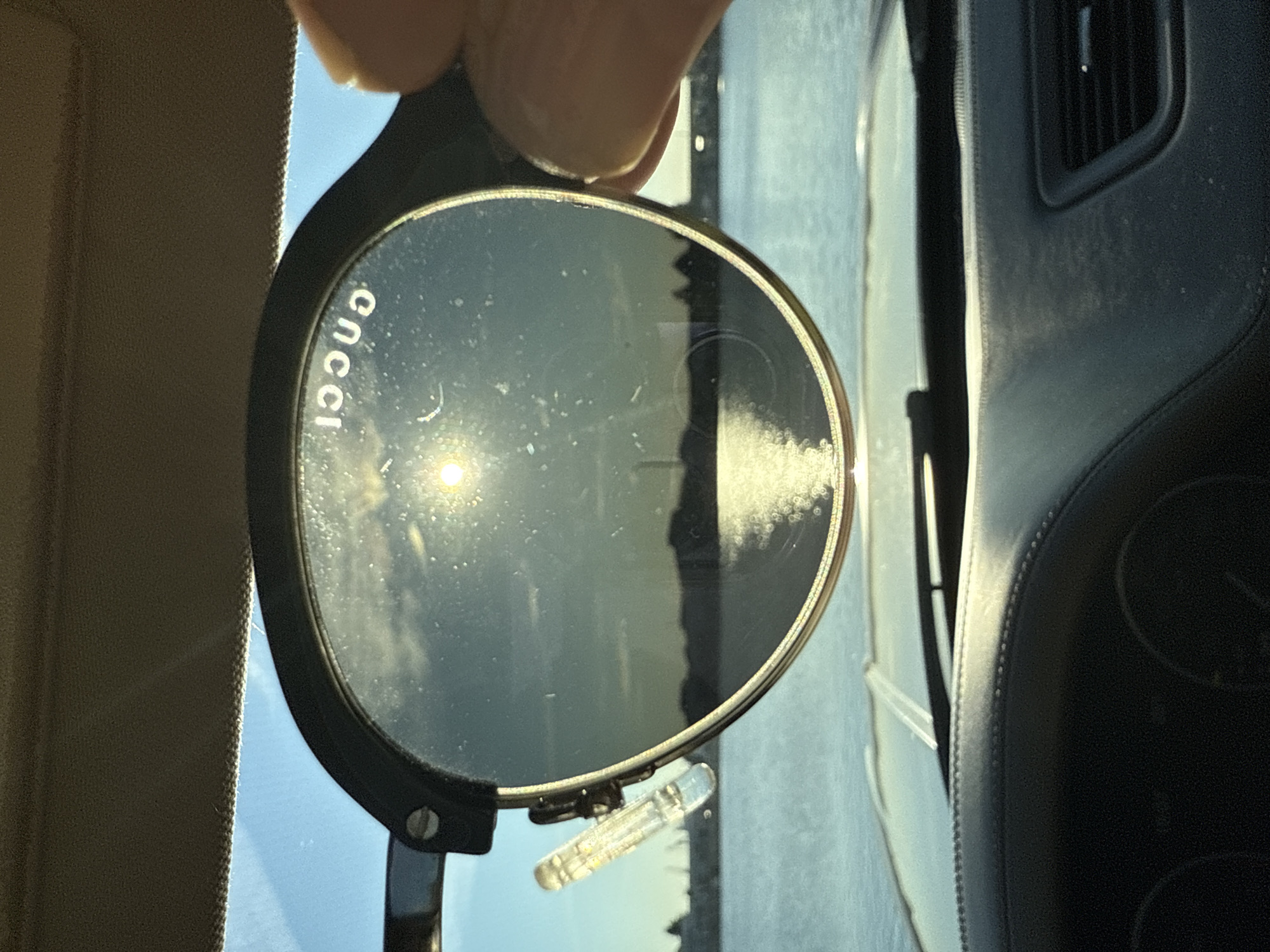 A pair of sunglasses is held up in front of a car's windshield, obscuring a view of a body of water with a faint city skyline in the distance. The sun is visible in the sky, reflecting off the water. The sunglasses have the Gucci logo etched onto the lens, which is reversed due to the reflection. The interior of the car, including the dashboard and air vents, is visible in the foreground. The image is taken during daylight, likely in the late afternoon due to the angle of the sun.