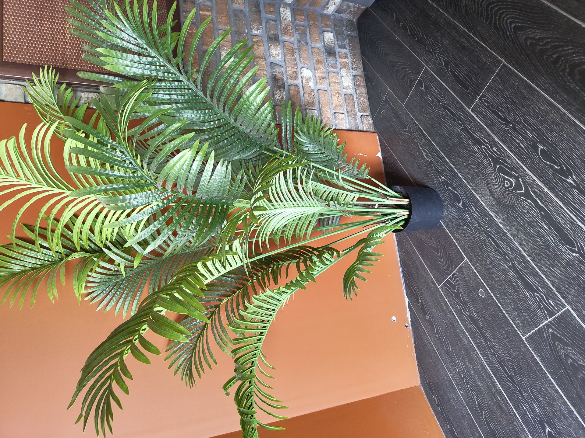 An artificial palm plant with numerous green fronds is positioned in a black pot on a dark wood floor. The plant is situated in a corner, with an orange wall to its left and a brick wall to its right. The brick wall appears to be made of small, rectangular bricks in varying shades of brown and gray. The floor is composed of dark, wide planks with a visible wood grain pattern and lighter lines between them.