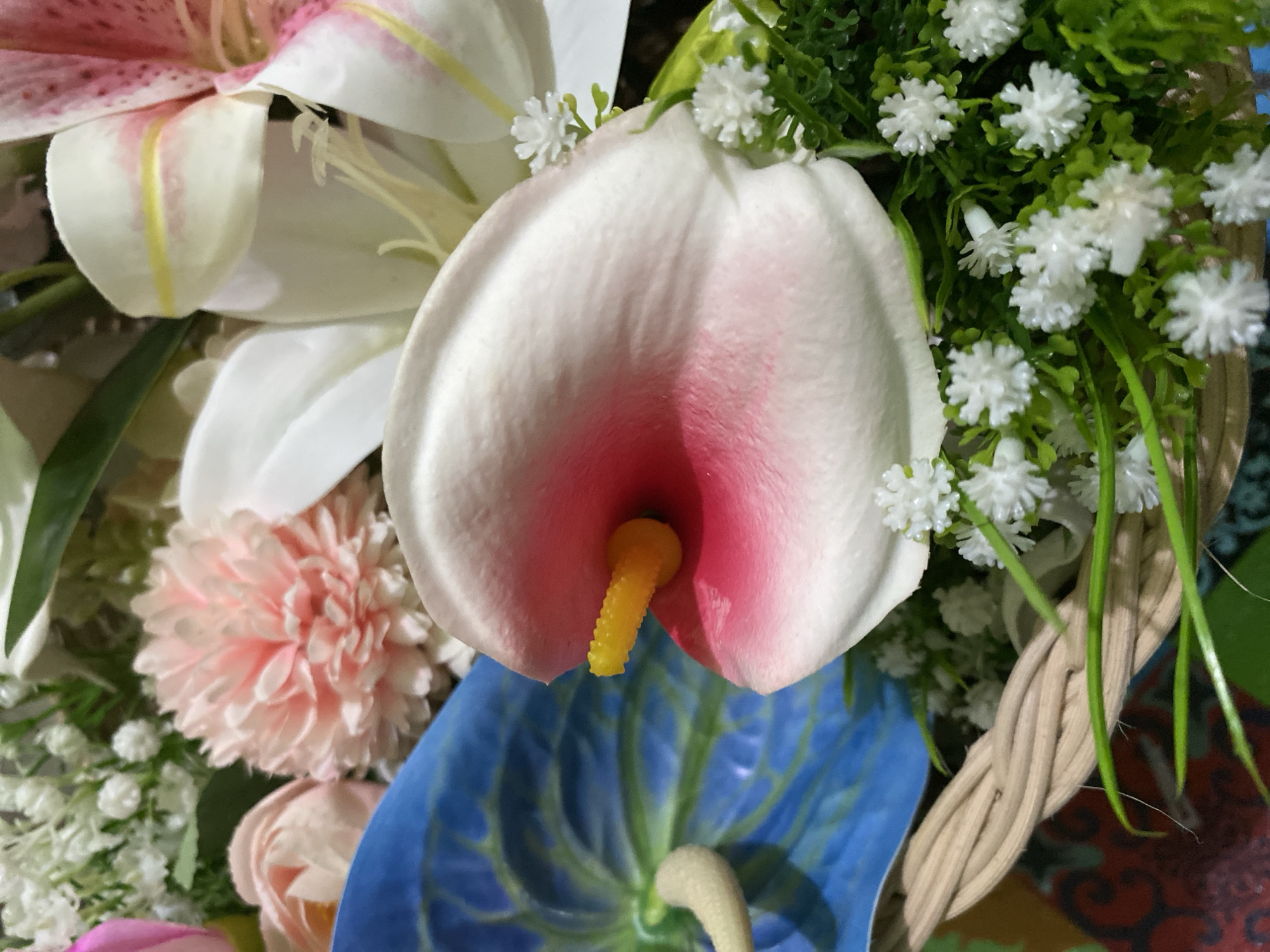 A close-up, still-life photograph captures a vibrant floral arrangement. The foreground is dominated by a white and pink calla lily, its bright yellow spadix prominently displayed. To the left, a striking blue anthurium flower adds a contrasting hue, while a cluster of delicate white flowers and green foliage fills the lower right portion of the frame. Hints of other flowers, including a pink carnation and soft pink roses, are visible in the background. The arrangement is partially nestled within a woven basket, with a colorful, patterned surface visible beneath. The scene is static, with no people or actions depicted. The photograph offers no information about its specific location within Kuala Pembuang, Indonesia, or any broader environmental context.