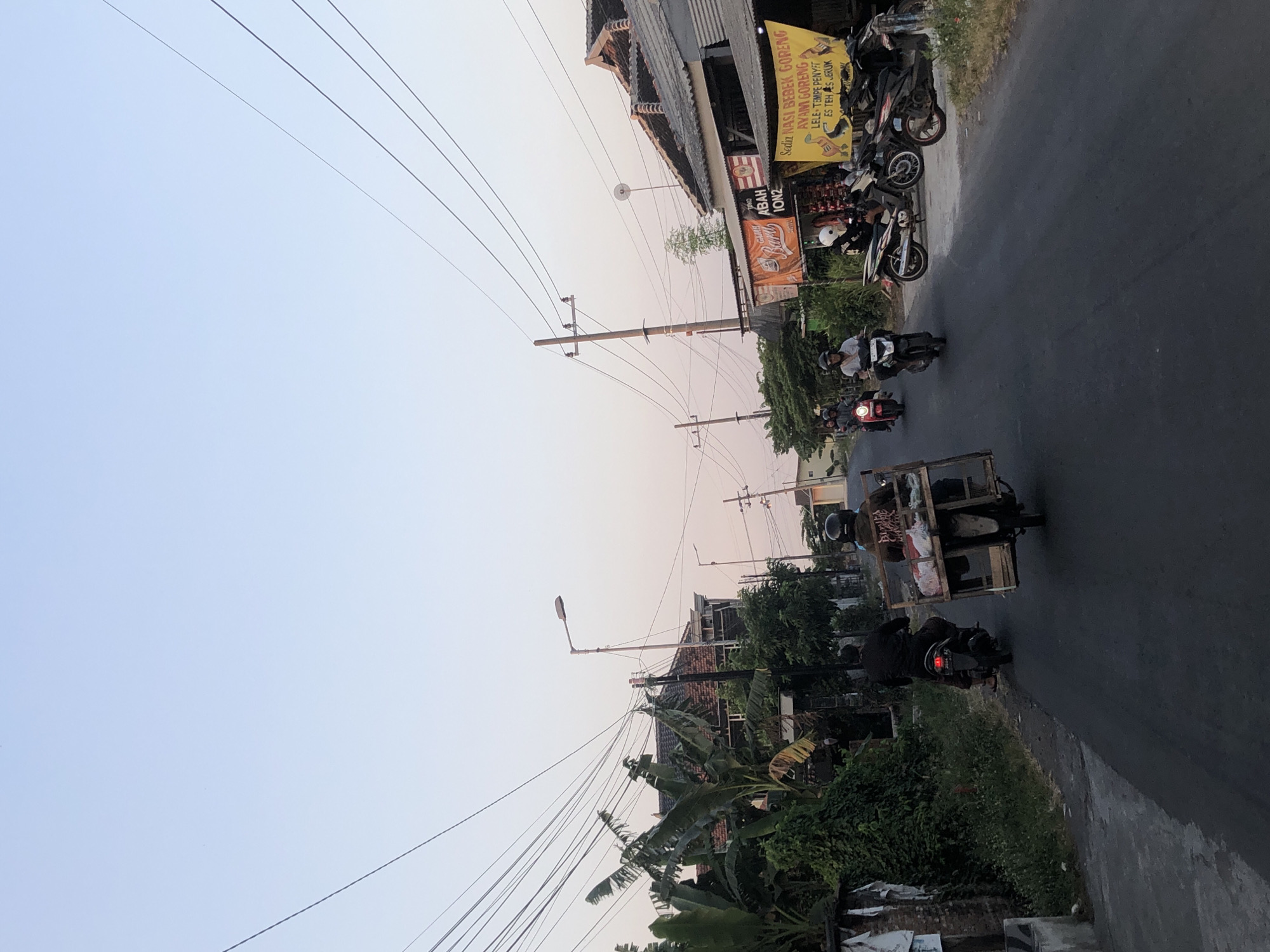 The image depicts a street scene in Glagah Arum, Indonesia, with a gentle twilight sky overhead. Several motorcycles are present on the paved road. In the foreground, a motorcycle is being ridden by an individual wearing a black jacket and helmet, carrying a wooden crate filled with items on the back. To the left, another motorcycle is occupied by a person in dark clothing. Further up the road, other motorcyclists are visible, some riding in groups. The sides of the street are lined with buildings, including structures with tiled roofs and houses with distinct architectural features. Vegetation, such as banana trees and other greenery, is interspersed throughout the scene. Additionally, various signs and banners are displayed, some with text indicating food vendors or businesses. Power lines crisscross above the street, supported by utility poles. The overall impression is of a typical evening in a residential or commercial area of Glagah Arum.