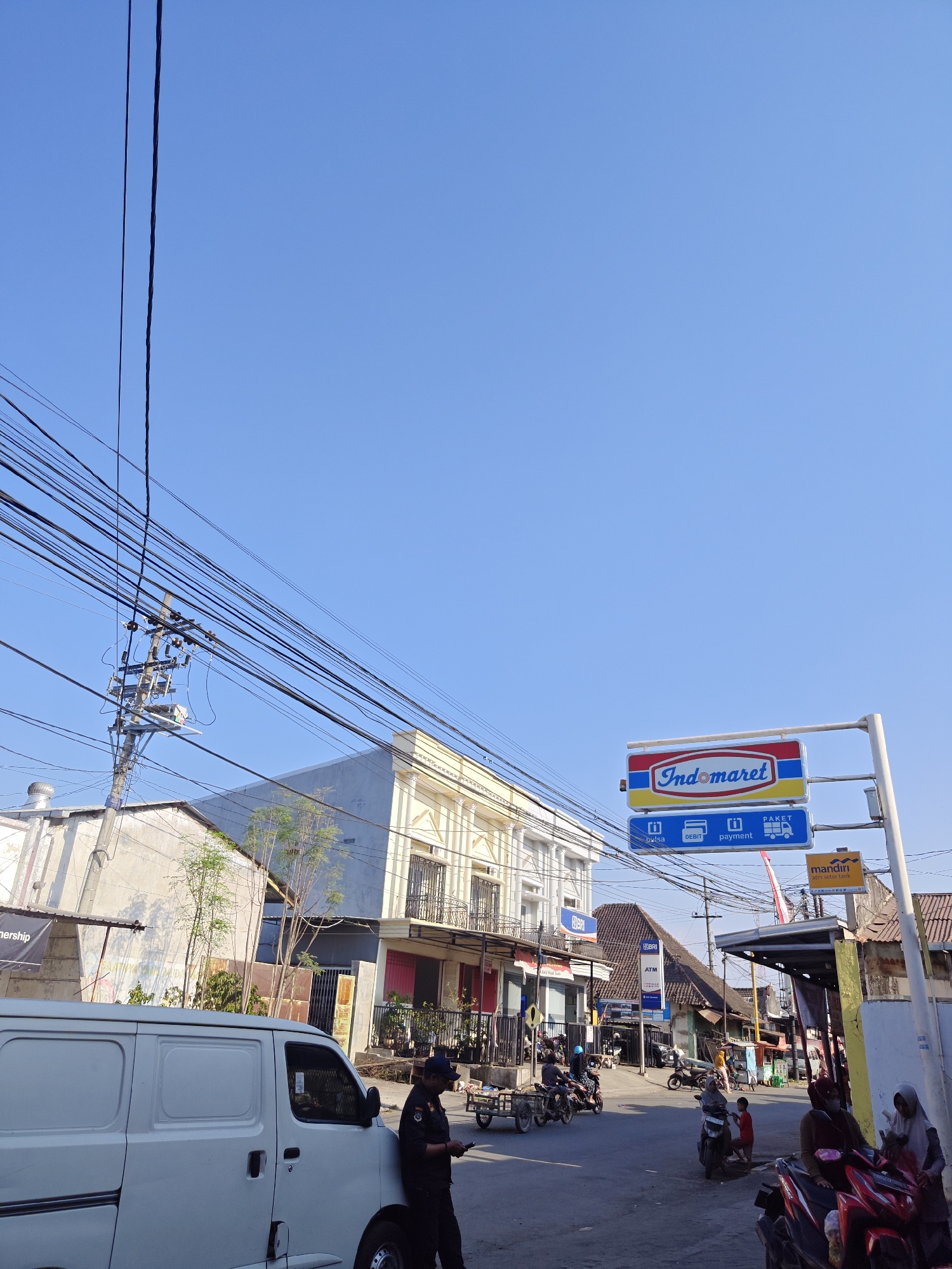 The image depicts a street scene in Klampokan, Indonesia, under a clear blue sky. Prominently featured is an Indomaret convenience store sign. In the foreground, a white van is parked on the right side of the road. A man in a dark uniform, possibly a security guard, is standing beside the van, looking at a mobile device. Several motorcycles are visible on the street, some with riders. Buildings line both sides of the street, including a light yellow multi-story building with balconies and a white building with exposed electrical wires and a utility pole. In the background, more buildings and a dense arrangement of overhead electrical wires dominate the view. The scene suggests a typical day in a residential or commercial area of the city.
