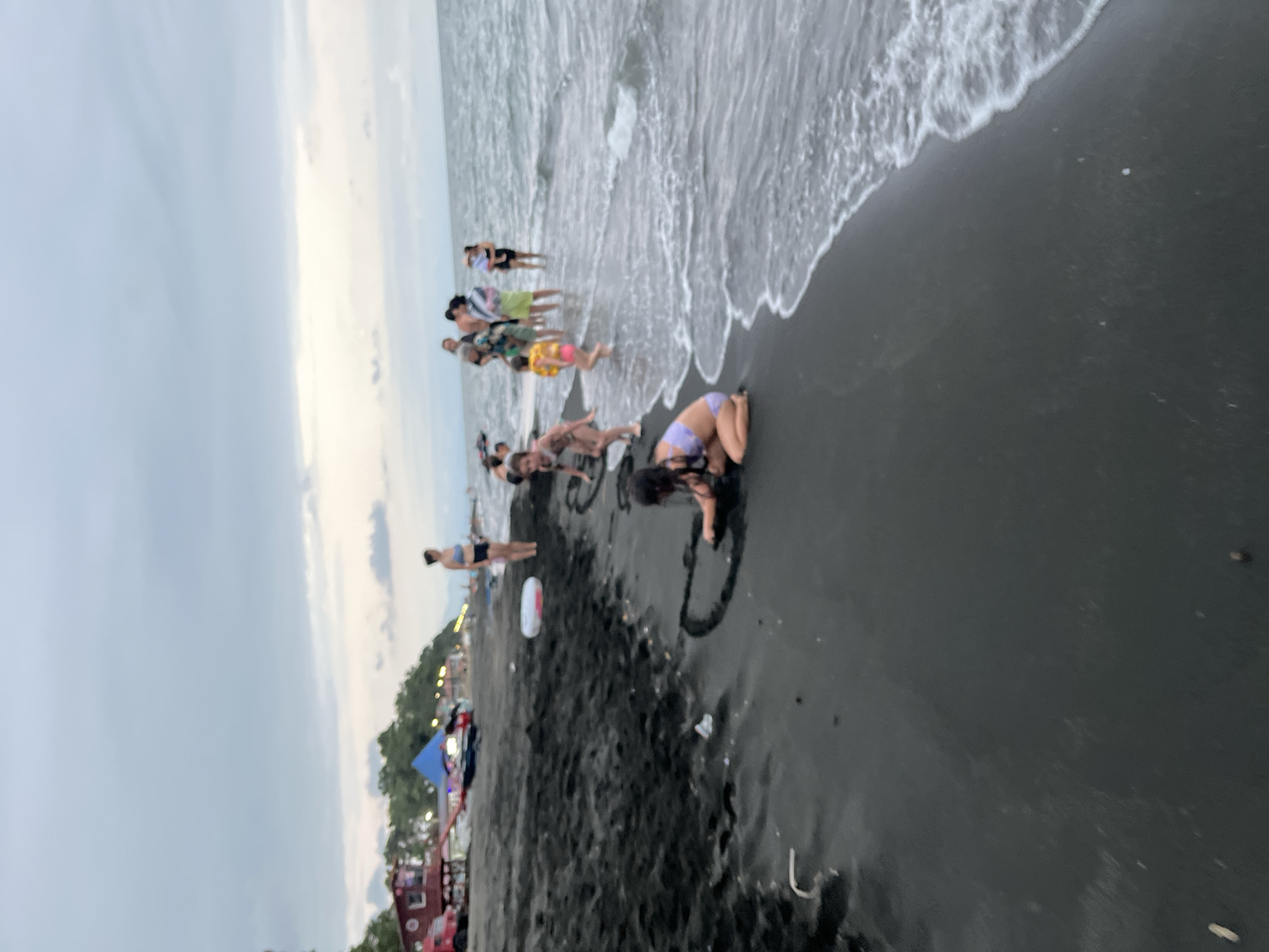 The image depicts a beach in Ureki, Georgia, with dark sand and the Black Sea. Several people are present, including adults and children, enjoying the water and the shore. In the foreground, a young girl in a purple swimsuit is kneeling on the wet sand, drawing a heart shape with her finger. Further back, a group of people, including children and adults, are standing in the shallow water, with some appearing to be playing. The background shows a line of trees and buildings along the coast, with a few tents and a red structure visible. The sky is overcast, suggesting a cloudy day.