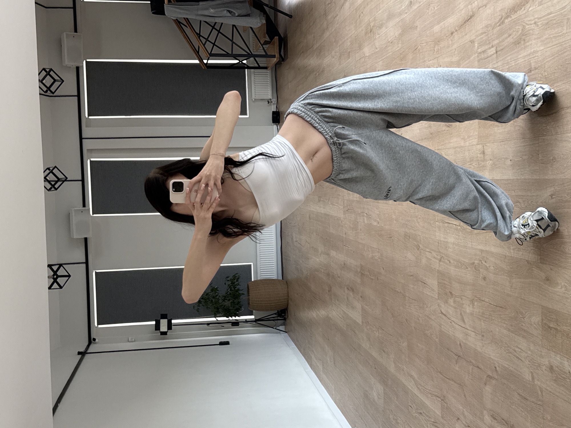 A young woman with dark hair is posing in a dance studio, holding a smartphone in her hands to take a mirror selfie. She is wearing a white crop top and loose grey sweatpants, with athletic shoes on her feet. The studio has a wooden floor, white walls, and large windows with grey roller blinds. The background shows a plant in a woven pot, a speaker, and a staircase with a railing. The lighting appears to be bright and natural. The overall scene suggests a practice or rehearsal space.