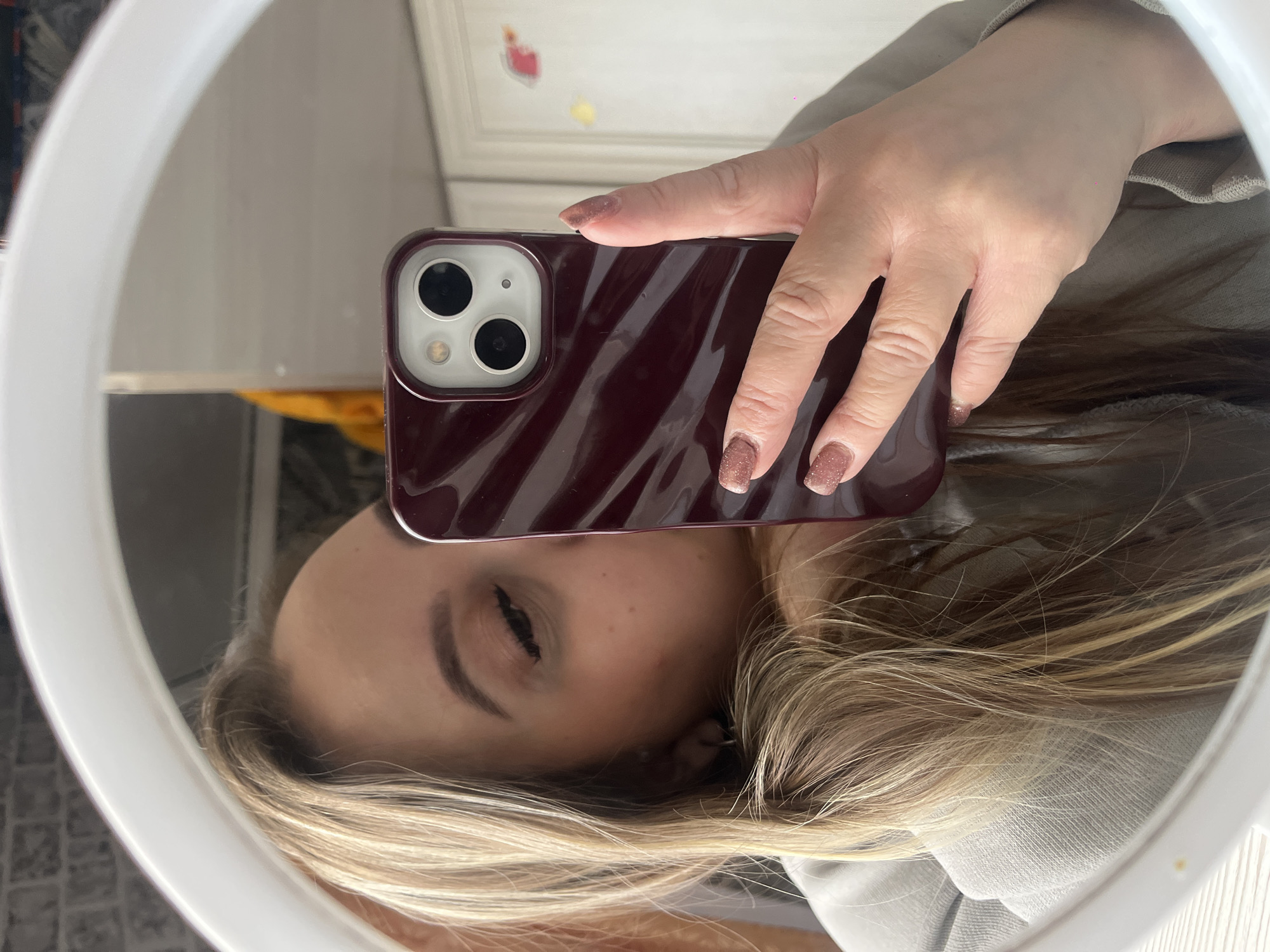 An indoor mirror selfie shows a person holding a smartphone, reflected in a circular or oval mirror with a white frame. The person's right hand, with glittery light brown or rose gold manicured nails, holds a dark red or burgundy smartphone case featuring a textured, wavy design and a dual-lens camera module with a flash.
The reflection reveals the upper part of the person's face beneath the phone, with blonde or light brown hair framing it. The eyes appear closed or are looking downwards. A gray or beige garment is visible on the reflected torso. The background reflected in the mirror behind the phone shows a light-colored wall, adorned with small, colorful decorations, including a red heart and a yellow shape. The lighting suggests it is daytime. No visible text is present in the image.