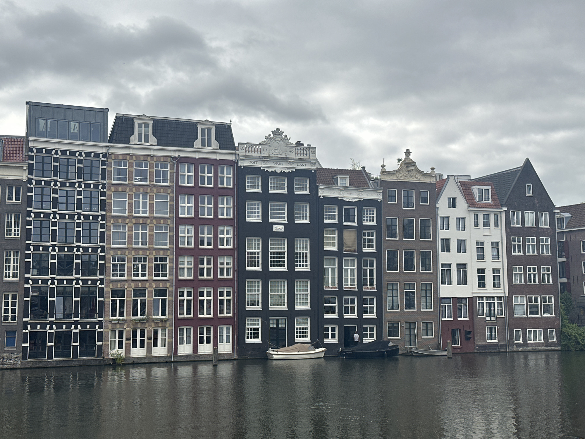 The image shows a row of canal houses in Amsterdam, Netherlands, under a cloudy sky. The buildings, of varying heights and architectural styles, are situated along a canal. The foreground is dominated by the dark, rippling water of the canal. Several boats are docked or moored along the canal bank. The buildings themselves exhibit a range of colors and materials, including brick, stucco, and modern glass facades. Windows are a prominent feature, with many buildings displaying numerous rectangular panes. Some buildings have decorative gables or cornices. The overall atmosphere is one of historical urban architecture meeting modern city life.