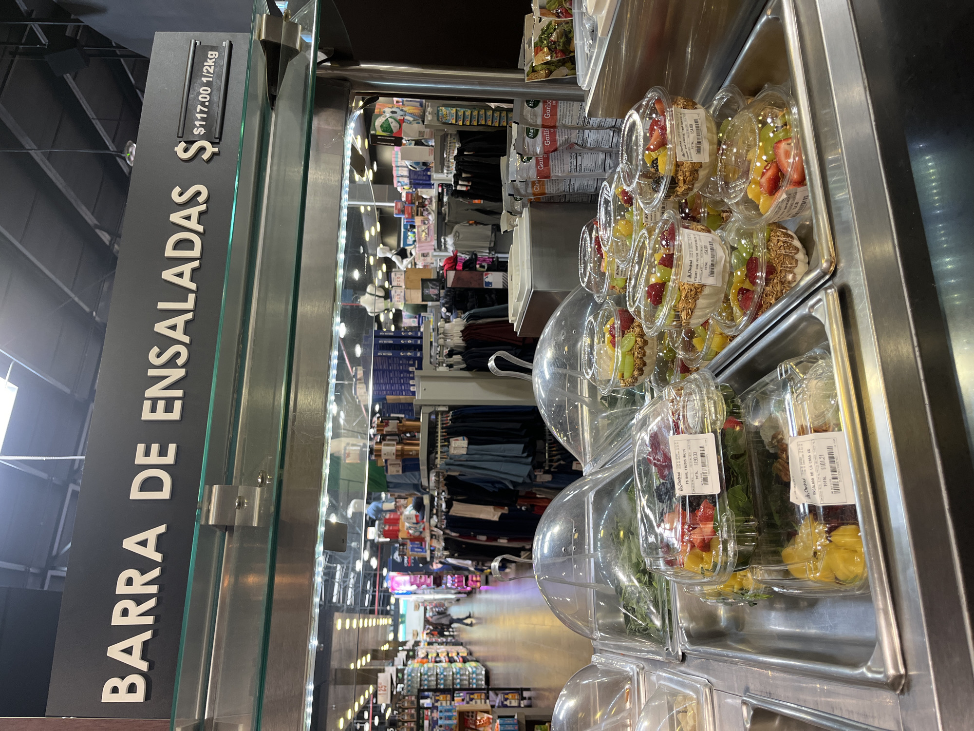 The image shows a salad bar, labeled "BARRA DE ENSALADAS" with a price of $117.00 per 1/2 kg. The salad bar is in a store with clothing and other merchandise visible in the background. The salads are pre-packaged in clear plastic containers, including salads with fruit, yogurt, and granola. One salad is labeled "FS BOAL FRUTOS ROJOS" with a total price of $190.00. The scene appears to be a modern supermarket in San Bernardino Tlaxcalancingo, Mexico.