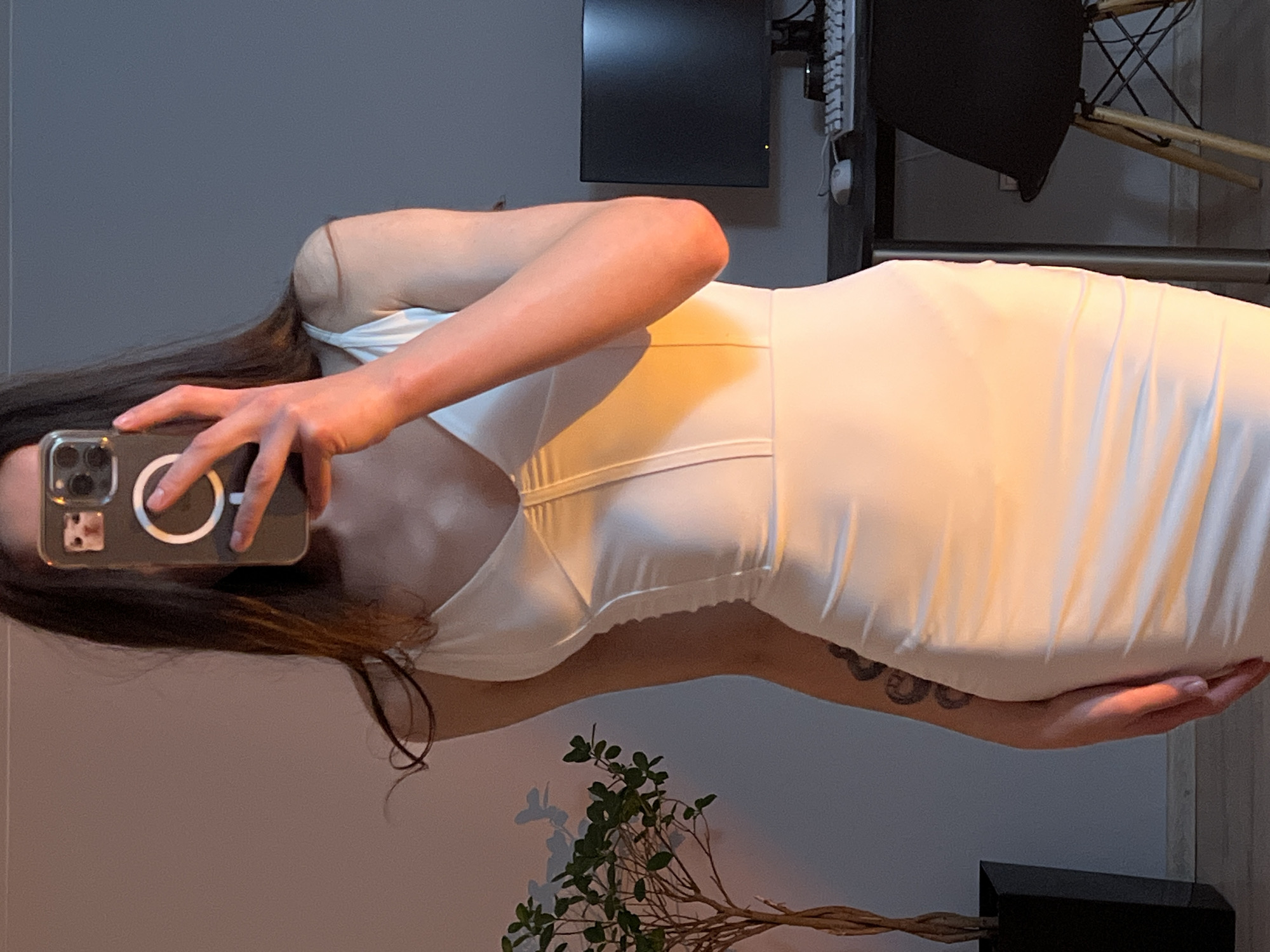 An indoor mirror selfie captures a person standing and posing in a light-colored, form-fitting dress. The individual, who has long dark hair, holds a smartphone (an iPhone with a visible MagSafe ring and clear case) to obscure their face while taking the photograph. A tattoo, possibly a coiled snake, is visible on their left forearm.
The setting appears to be a room within a residence, possibly in Ternopil, Ukraine. In the background, a dark monitor or television screen is mounted on the wall, with a keyboard and computer mouse visible adjacent to it. Part of a chair with wooden legs and a dark seat can be seen behind the person. A metallic horizontal bar or rail crosses the mid-ground. Towards the bottom of the image, a potted plant with green leaves and a twisted stem adds a touch of nature to the scene. The walls are a neutral tone, and artificial lighting casts warm highlights on the dress, suggesting an indoor ambiance. No discernible text is visible in the image.